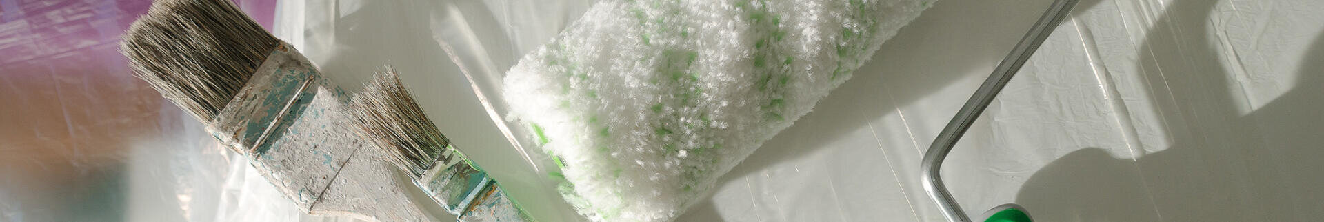 Nahaufnahme eines Pinsels und einer flauschigen Farbrolle, die auf einer weißen Fläche liegen und auf Vorbereitungen für Maler- oder Heimwerkerarbeiten hindeuten.