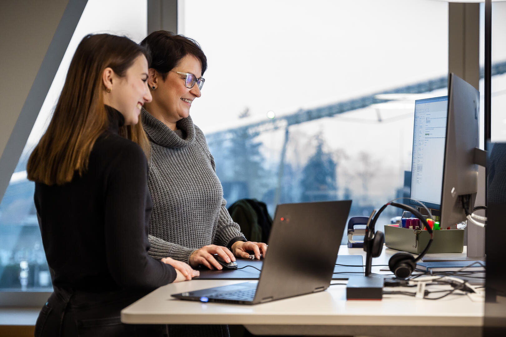 Zwei Frauen, die lächelnd an einem Stehpult mit Laptops und einem Monitor in einem modernen Büro arbeiten, neben großen Fenstern mit einem verschwommenen Blick nach draußen.