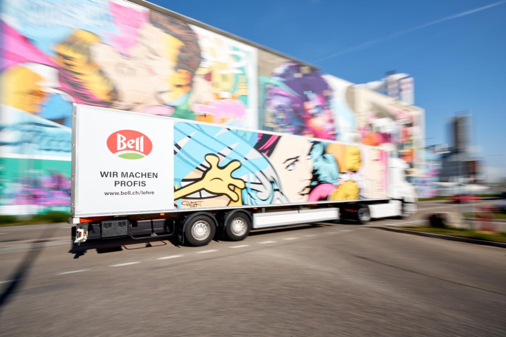 Ein Lastwagen mit farbenfrohen Pop-Art-Grafiken und dem Bell-Logo fährt an einem sonnigen Tag an einem Gebäude mit bunten Wandmalereien vorbei.