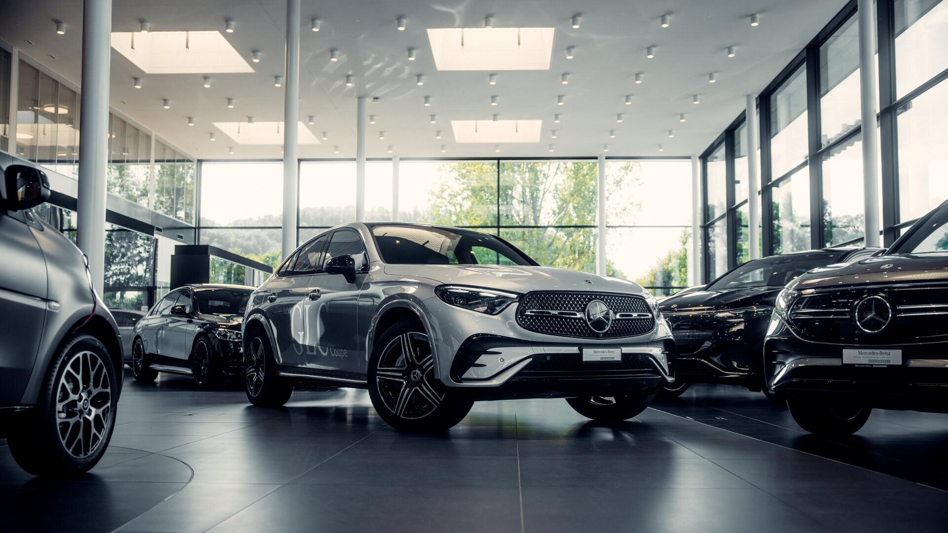 Ein silberner Mercedes-Benz-SUV steht in einem modernen Autohaus mit großen Fenstern, umgeben von anderen Autos und heller Deckenbeleuchtung.