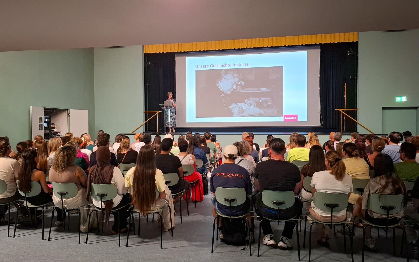 Eine Frau spricht auf der Bühne zu einem Publikum, das in Reihen sitzt; hinter ihr ist auf einer großen Leinwand ein Schwarz-Weiß-Foto zu sehen, auf dem der deutsche Text „Unsere Geschichte in Kürze“ steht.