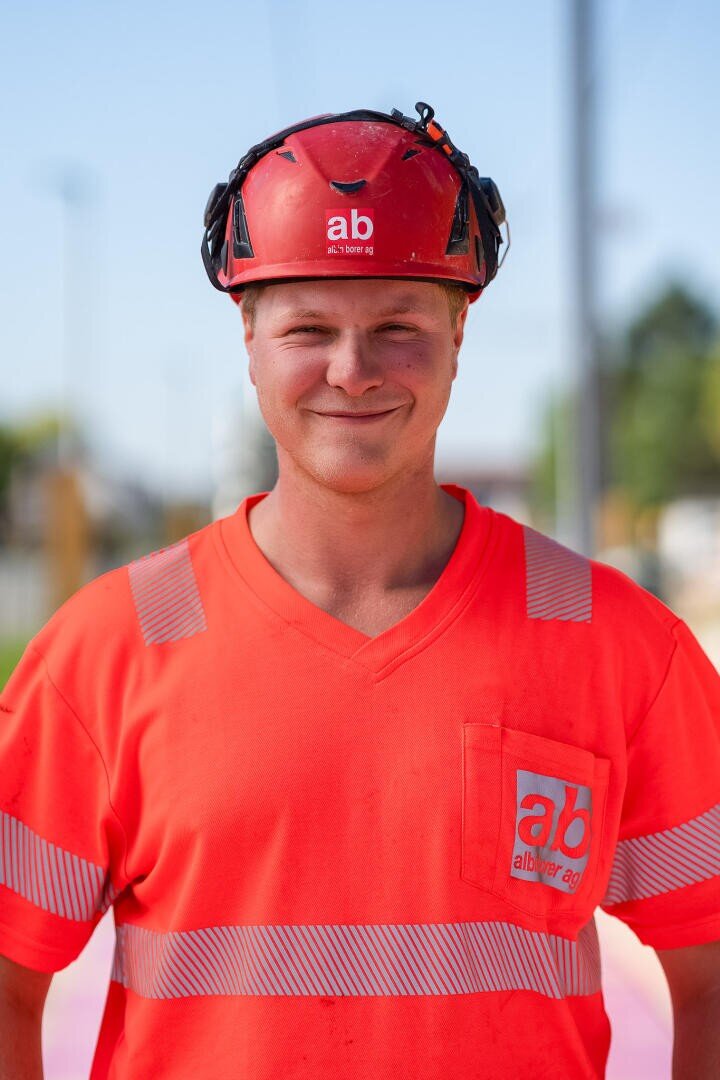 Ein Bauarbeiter trägt einen roten Schutzhelm und ein orangefarbenes Sicherheitshemd mit einem „ab“-Logo. Er steht im Freien und lächelt in die Kamera.