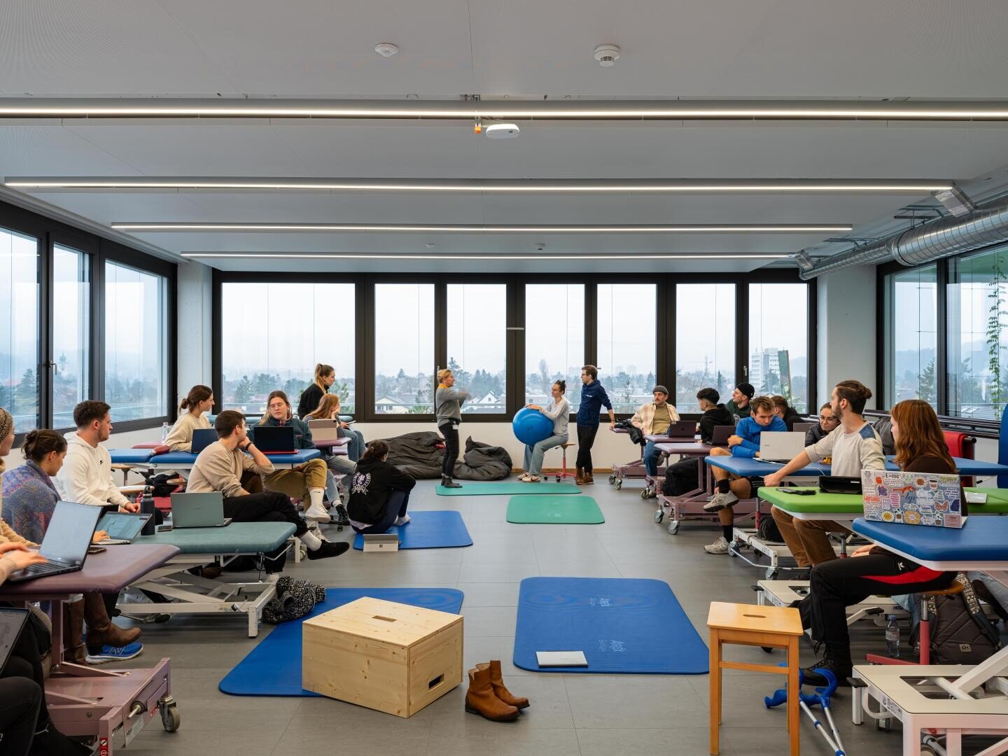 Ein Klassenzimmer, in dem Schüler an Tischen und auf Trainingsmatten arbeiten. Einige nutzen Laptops, während andere zu zweit miteinander arbeiten. Durch große Fenster hat man einen Blick auf die Stadt. Auf dem Boden stehen eine Holzkiste und Schuhe. Es herrscht eine Atmosphäre der Zusammenarbeit.