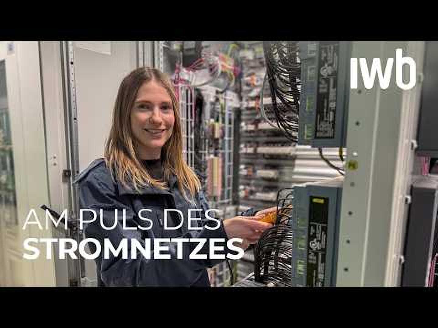 Eine Frau steht lächelnd vor Schalttafeln und hält Kabel in der Hand. Der Text auf dem Bild lautet „IWB“ und „Am Puls des Stromnetzes“.