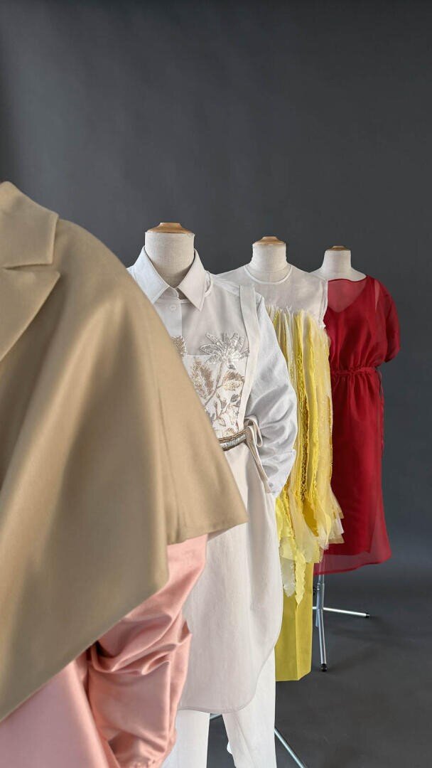 Vier Schaufensterpuppen zeigen stilvolle Outfits: einen beigen Mantel mit rosafarbenen Details, ein weißes besticktes Hemd mit Hose, ein gelbes Rüschenkleid und ein rotes Kleid, alles vor einem dunkelgrauen Hintergrund.