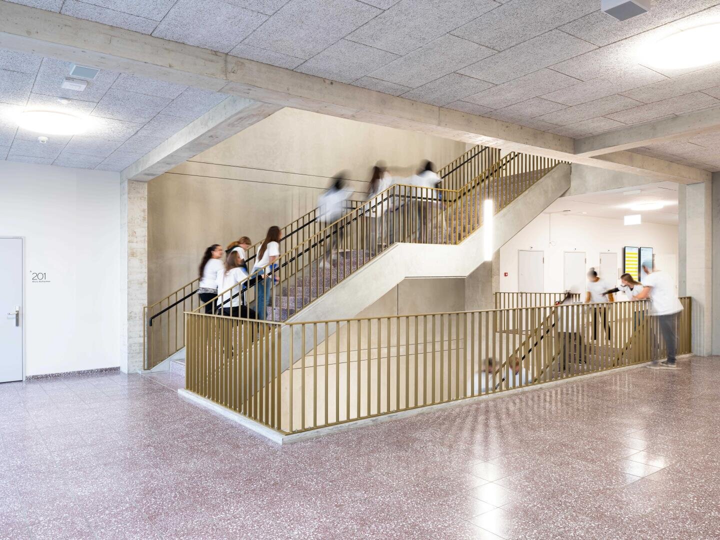 Schüler in weißen Hemden gehen in einem hellen Schulflur eine moderne Innentreppe mit goldenen Geländern auf und ab; einige von ihnen wirken aufgrund der Bewegung verschwommen.