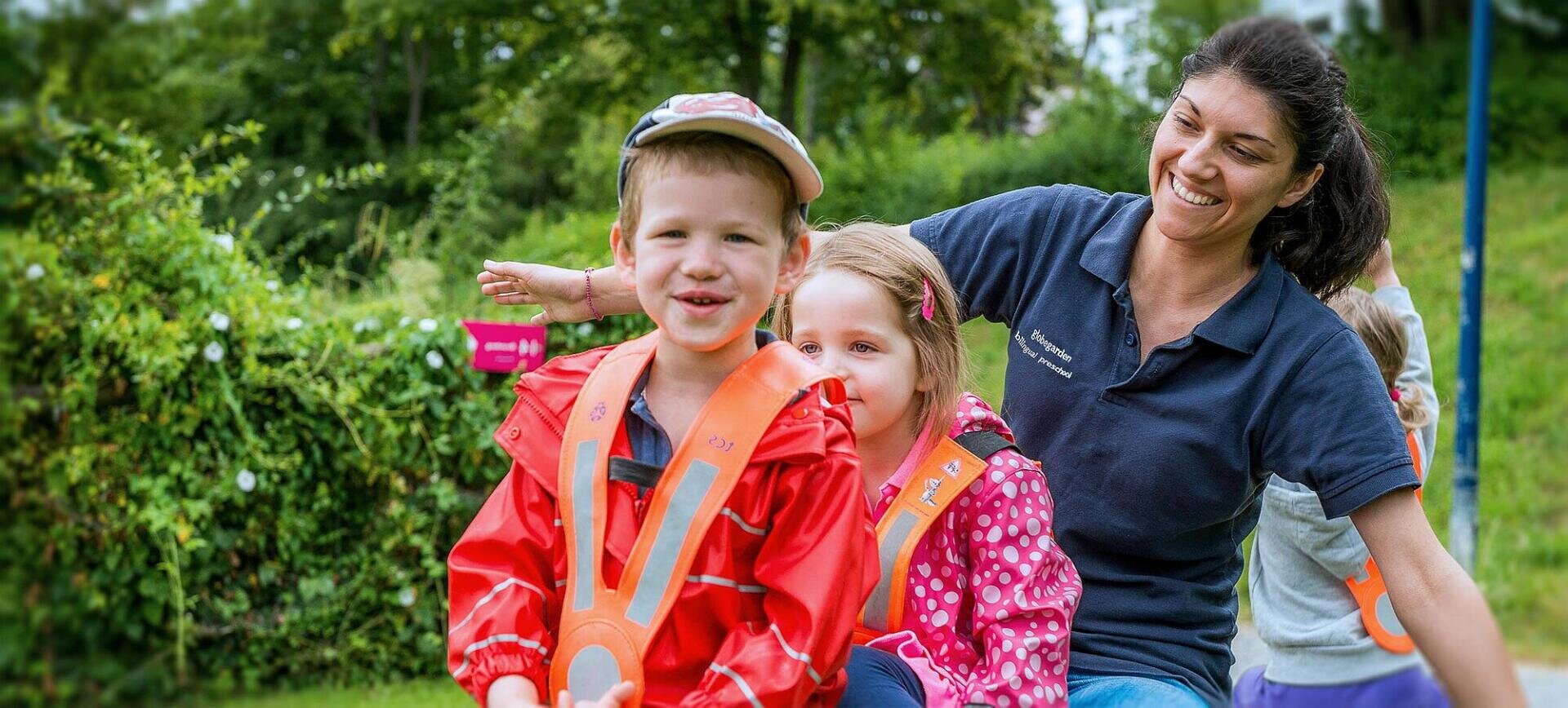 Eine lächelnde Frau und drei kleine Kinder, die alle bunte Jacken tragen, sitzen in einer Reihe im Freien. Die Kinder sehen glücklich aus, und der Hintergrund zeigt Grün und Bäume.