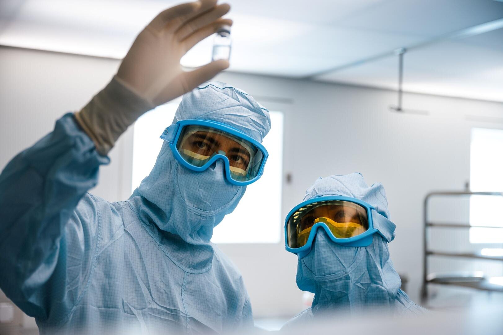 Zwei Wissenschaftler in blauen Schutzanzügen und mit Schutzbrillen untersuchen ein Fläschchen in einer hellen, sterilen Laborumgebung.