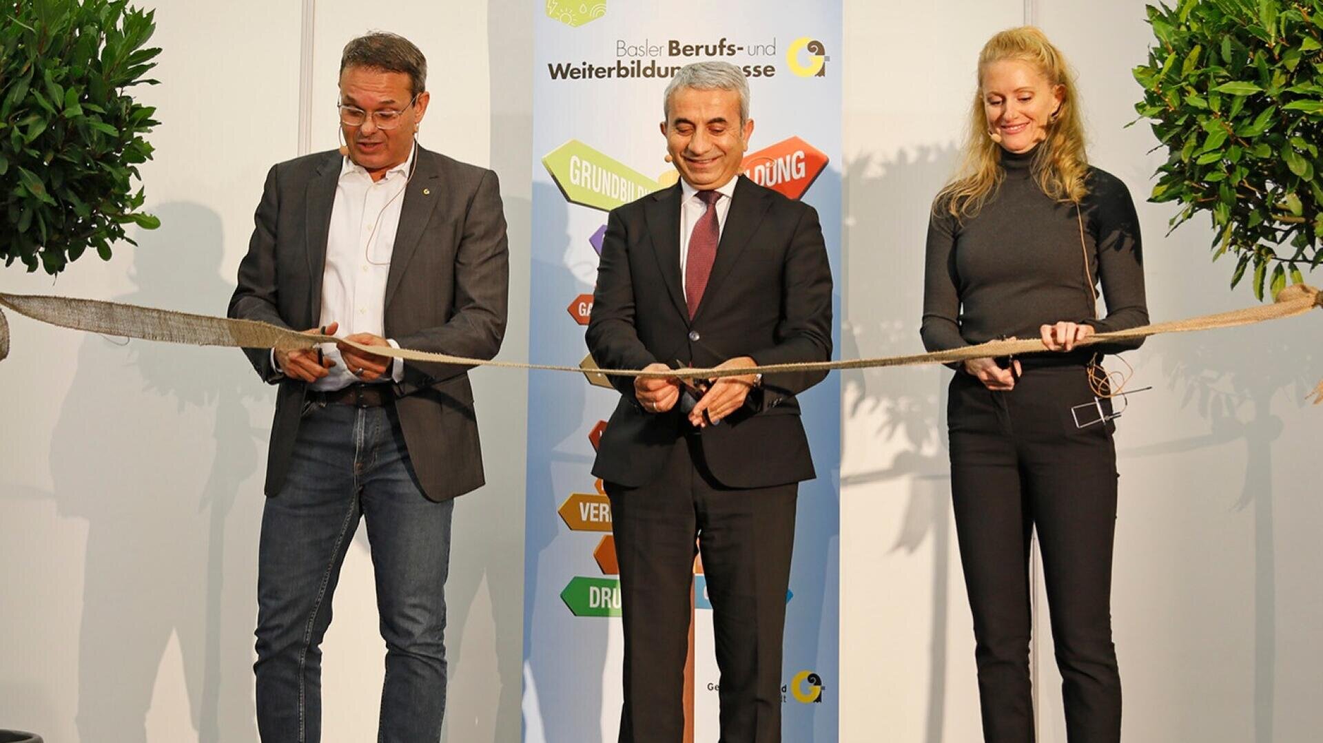 Drei Personen schneiden lächelnd gemeinsam ein langes Band bei einer Veranstaltung durch und stehen vor einem Schild mit bunten Pfeilen und Text. Zwei Topfpflanzen sind auf beiden Seiten der Gruppe zu sehen.