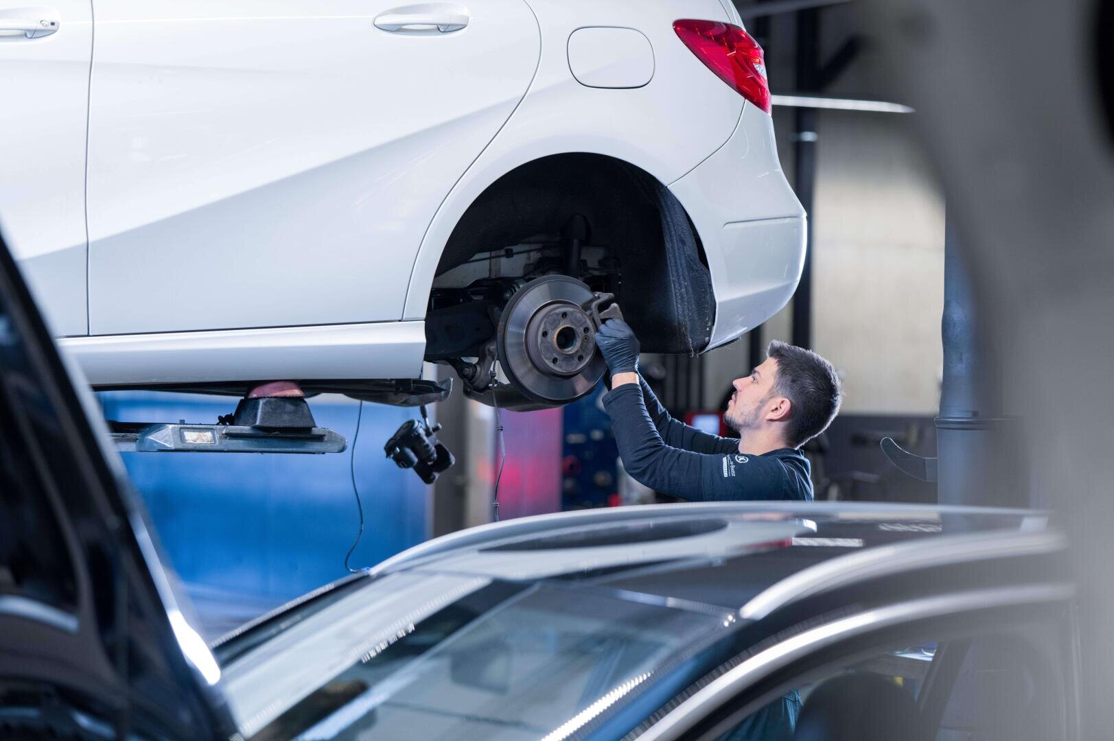 Ein Mechaniker untersucht den Bereich des Hinterrads eines weißen Autos, das in einer Autowerkstatt auf einer Hebebühne steht.