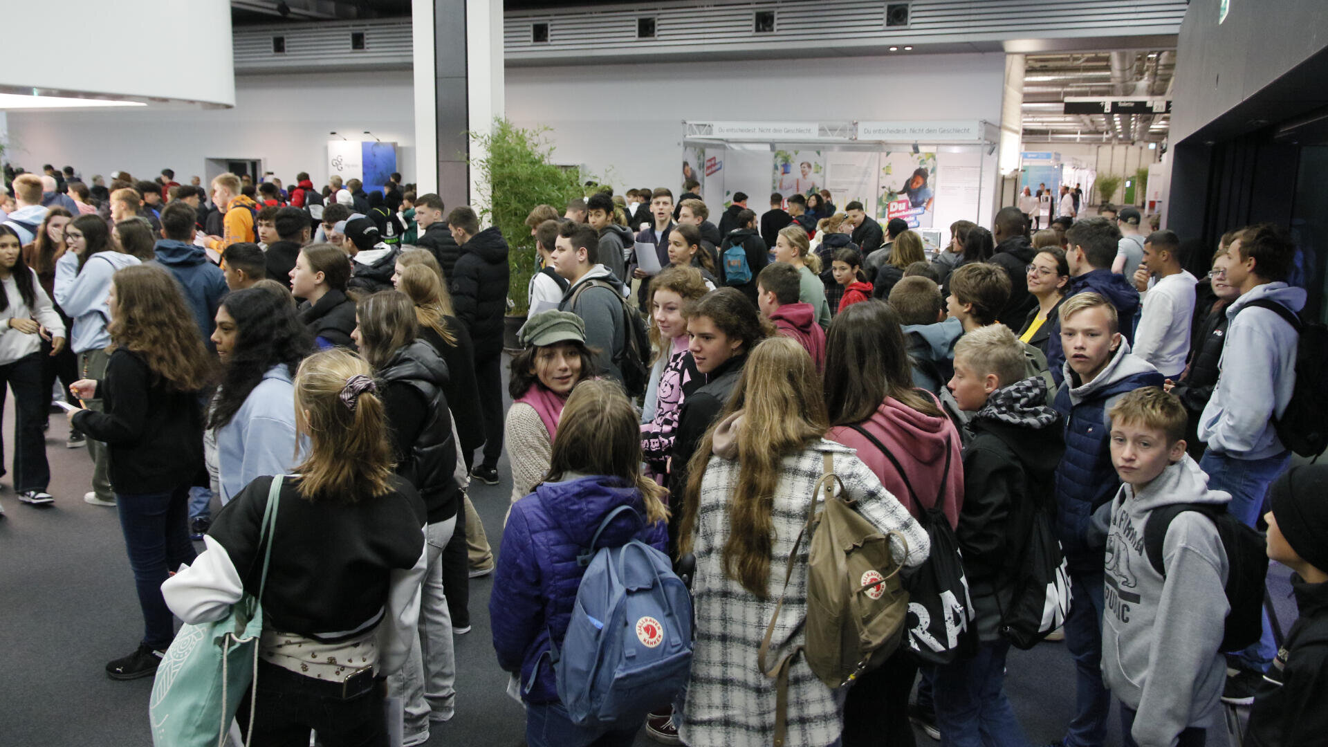 Eine große Gruppe von Schülern, viele mit Rucksäcken und Jacken, versammelt sich in einem belebten, modernen Flur oder einer Lobby, unterhält sich und wartet. Einige schauen in die Kamera, andere schauen sich um oder unterhalten sich mit Freunden.