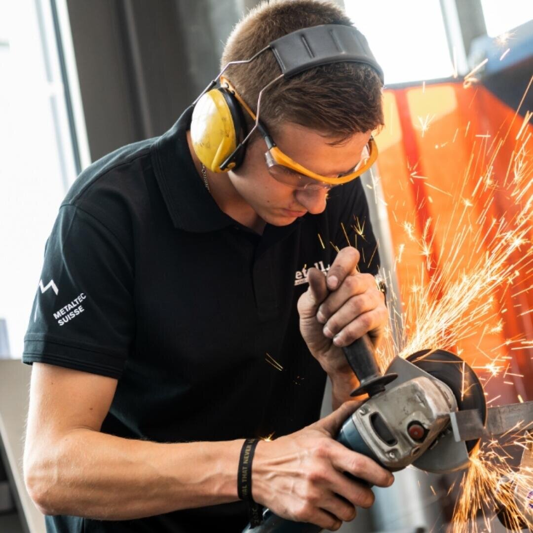 Eine Person, die eine Schutzbrille und einen Gehörschutz trägt, bedient einen Winkelschleifer und erzeugt dabei helle Funken, während sie in einer industriellen Umgebung an einem Metallgegenstand arbeitet.