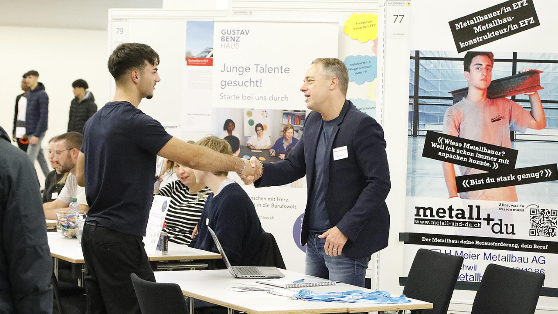 Zwei Männer schütteln sich am Stand einer Berufsmesse die Hände. Der eine trägt dunkle Kleidung, der andere einen Blazer. Im Hintergrund sitzen weitere Personen an Tischen. Auf Plakaten und Bannern werden Stellenangebote und Unternehmen angepriesen.