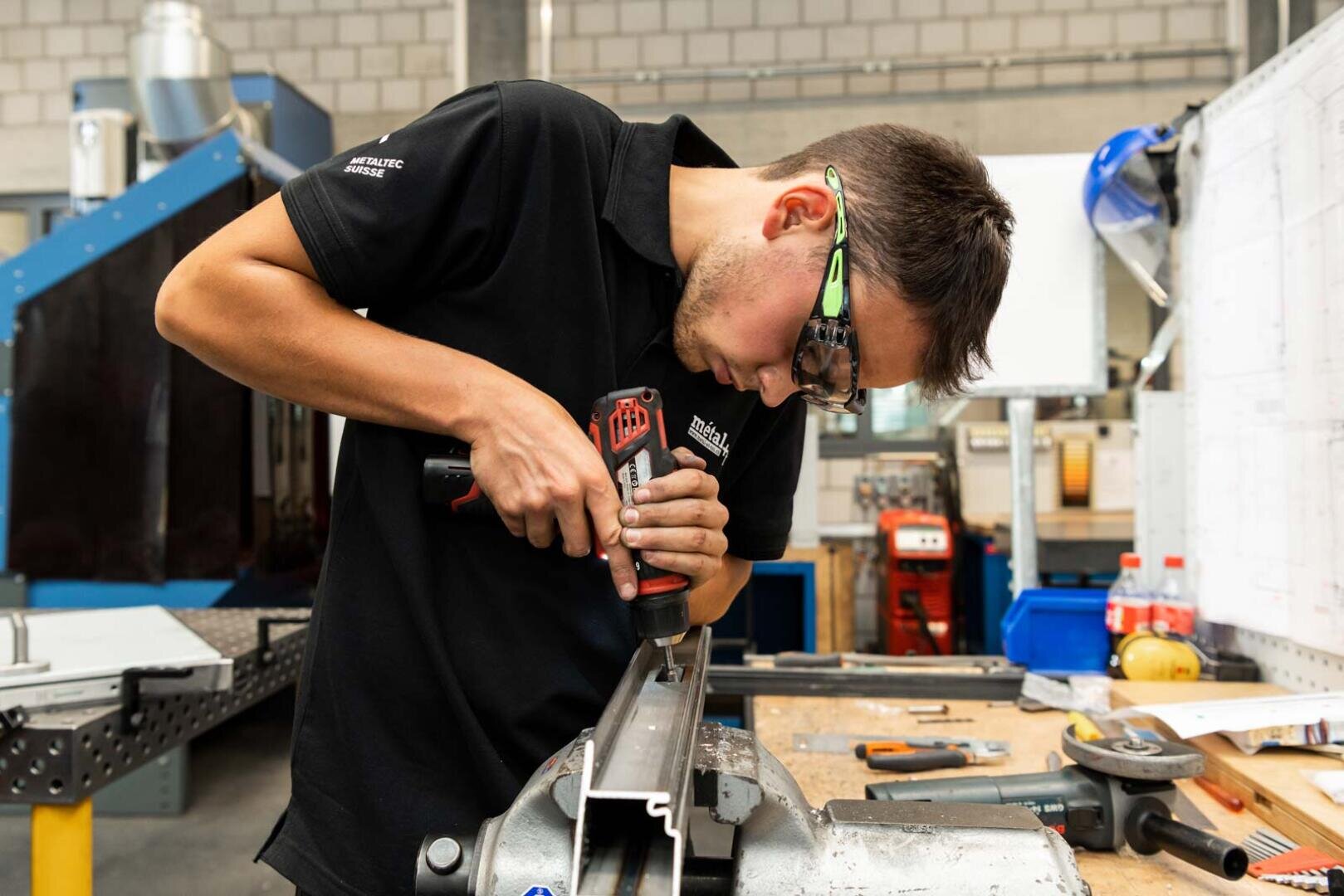 Eine Person, die eine Schutzbrille trägt, bearbeitet mit einer Bohrmaschine ein Metallstück, das auf einer Werkbank in einer Industriewerkstatt eingespannt ist. Auf dem Tisch und im Hintergrund sind Werkzeuge und Geräte zu sehen.
