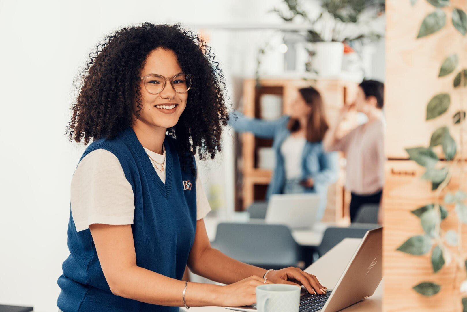 Eine lächelnde Person mit lockigem Haar und Brille tippt an einem Laptop an einem Schreibtisch. Im verschwommenen Hintergrund unterhalten sich zwei Personen in einem modernen, hellen Büroraum mit Pflanzen.