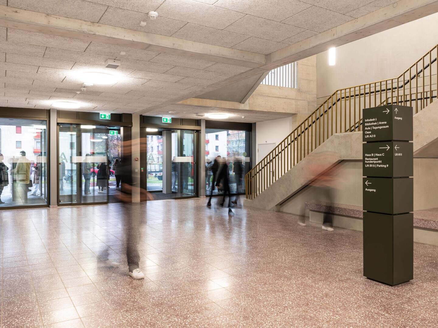 Moderne Gebäude-Lobby mit Terrazzoboden, Treppe mit goldenem Geländer, großen Fenstern und vorbeigehenden Menschen. In der Mitte befindet sich ein schwarzes Wegweiser-Schild. Einige Personen sind unscharf abgebildet, was auf Bewegung hindeutet.