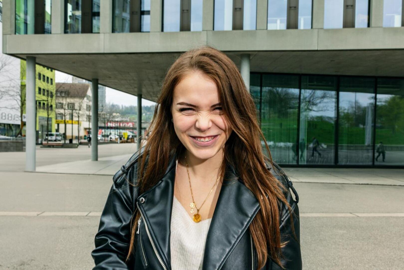 Eine junge Frau mit langen braunen Haaren, die eine schwarze Lederjacke und ein weißes Oberteil trägt, lächelt an einem bewölkten Tag im Freien vor einem modernen Glasgebäude.