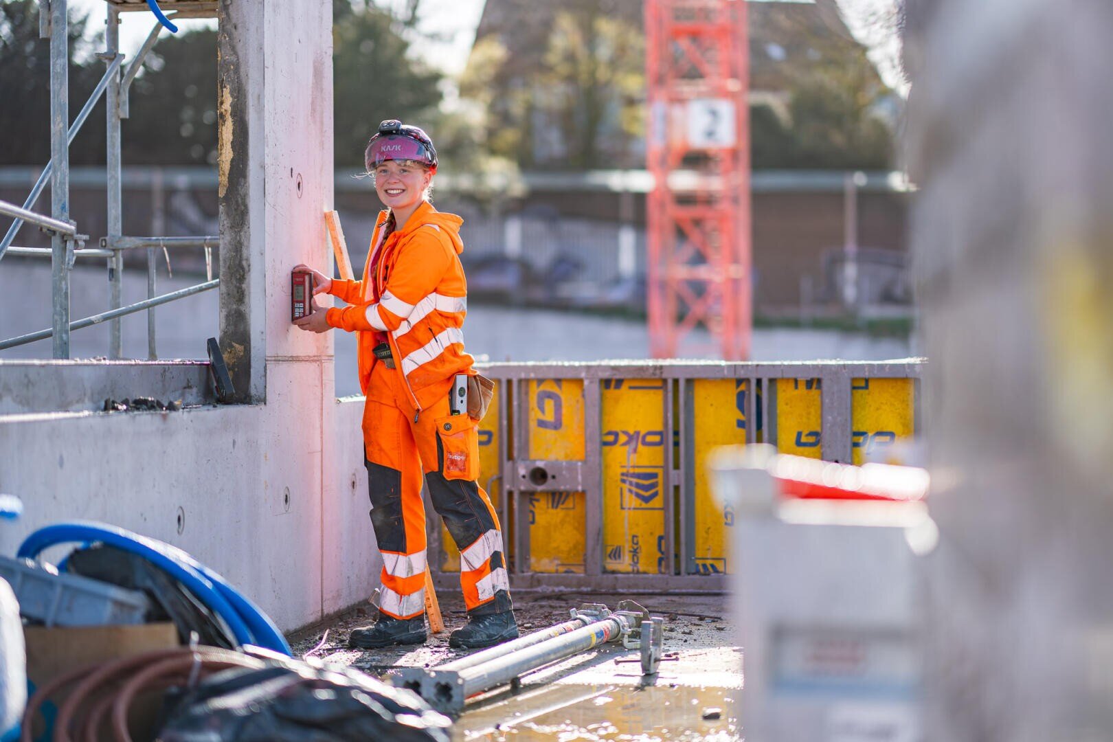 Ein Bauarbeiter in orangefarbener Schutzkleidung und mit einem Helm lächelt, während er auf einer Baustelle arbeitet, umgeben von Betonwänden, Gerüsten und Baumaterialien.