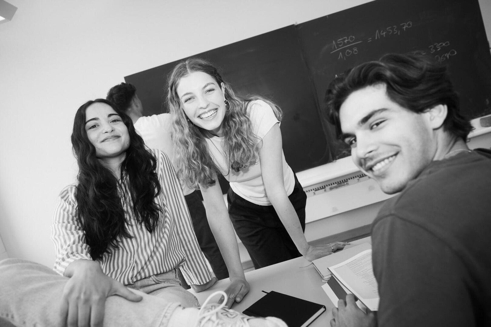 Drei junge Erwachsene, zwei Frauen und ein Mann, sitzen in einem Klassenzimmer mit einer Kreidetafel im Hintergrund und lächeln in die Kamera. Eine Frau lehnt sich an ein Pult, und man sieht Bücher und Hefte.