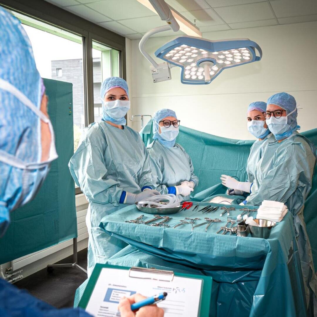 Vier Chirurgen in OP-Kleidung und Masken stehen mit chirurgischen Instrumenten um einen Operationstisch herum und blicken auf eine Person im Vordergrund, die ein Klemmbrett in der Hand hält. Im Raum sind helle Operationslampen und ein Fenster zu sehen.