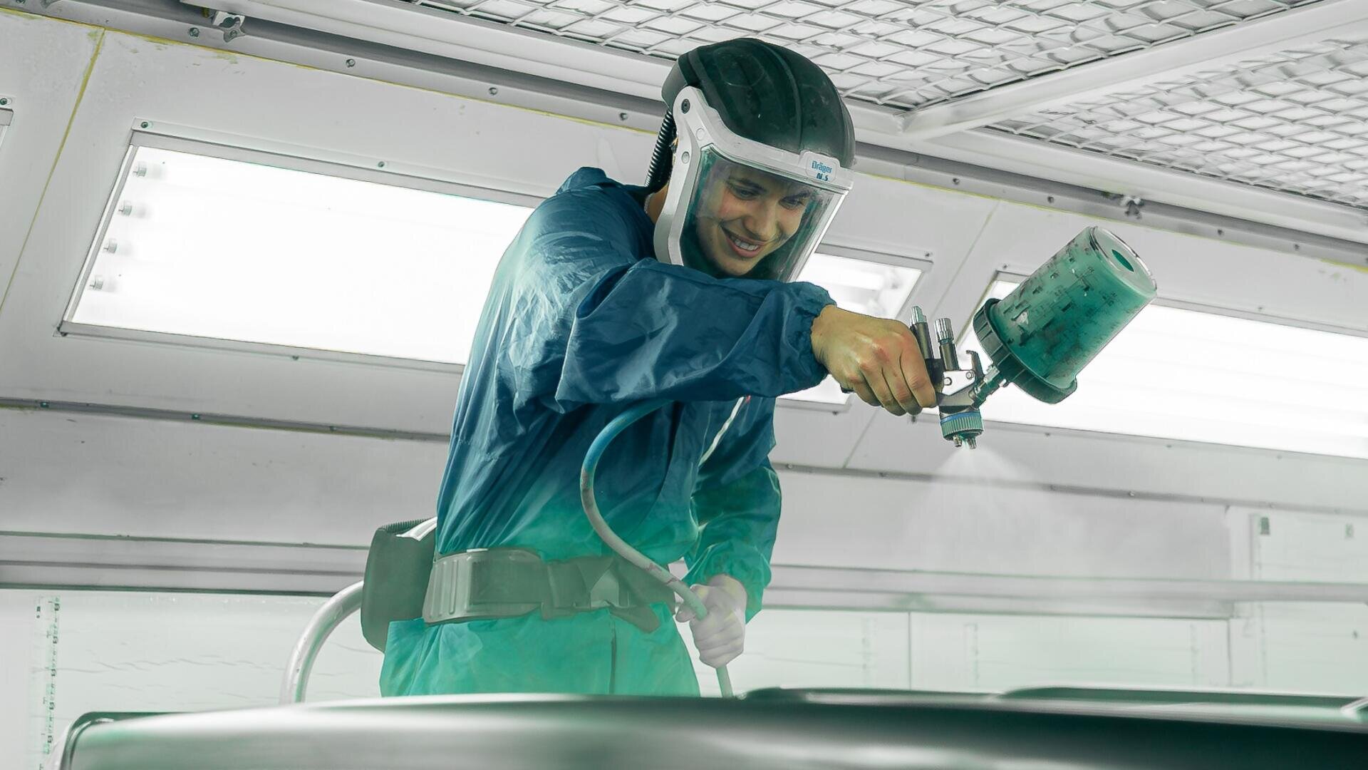 Eine Person in Schutzkleidung lackiert oder beschichtet mit einer Spritzpistole eine Oberfläche in einer gut beleuchteten Industriekabine.