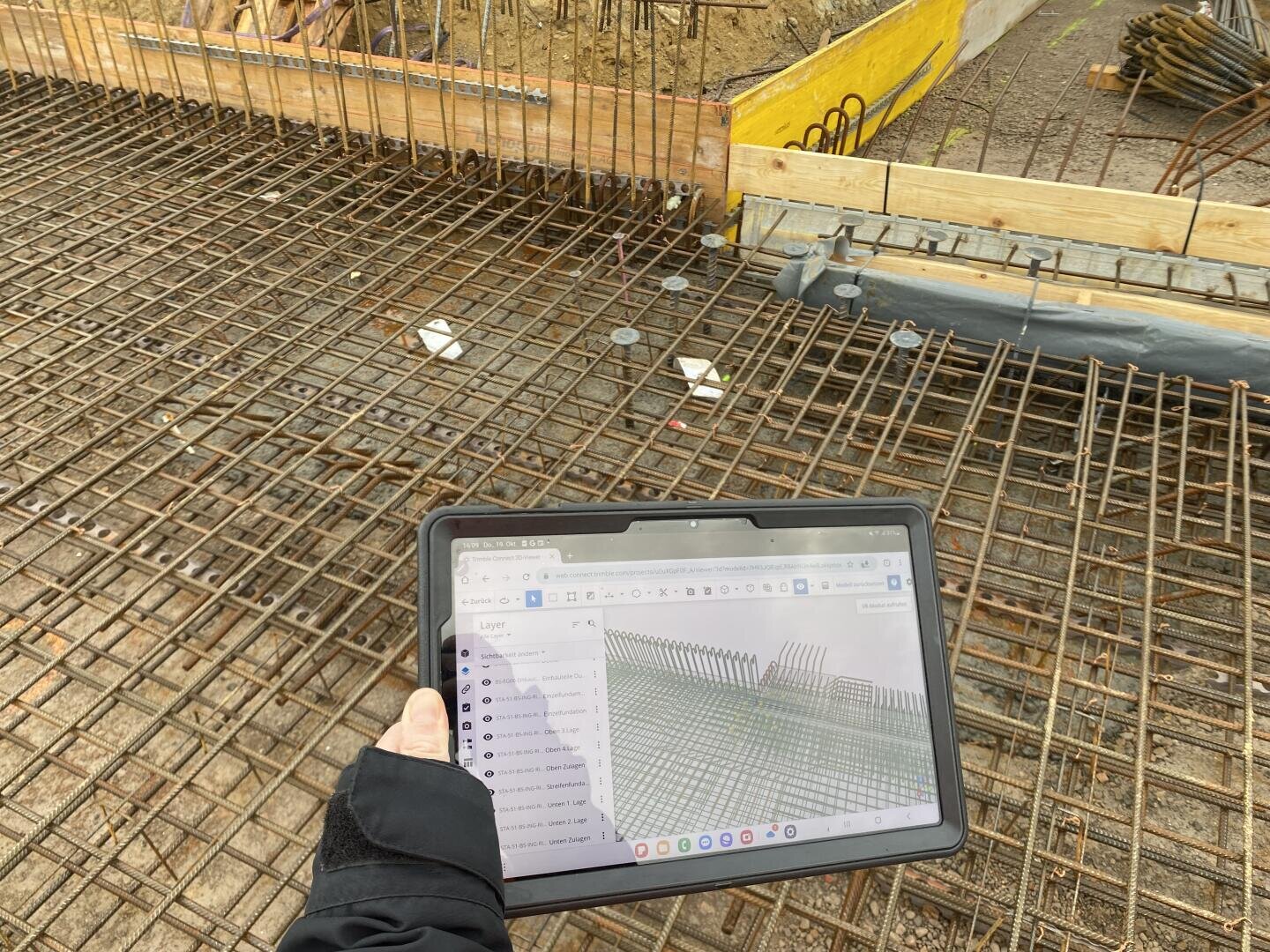 Eine Person hält auf einer Baustelle ein Tablet in der Hand, auf dem ein 3D-Bauplan über einem Geflecht aus Stahlbewehrungsstäben angezeigt wird; im Hintergrund sind Holzschalungen und freiliegender Boden zu sehen.