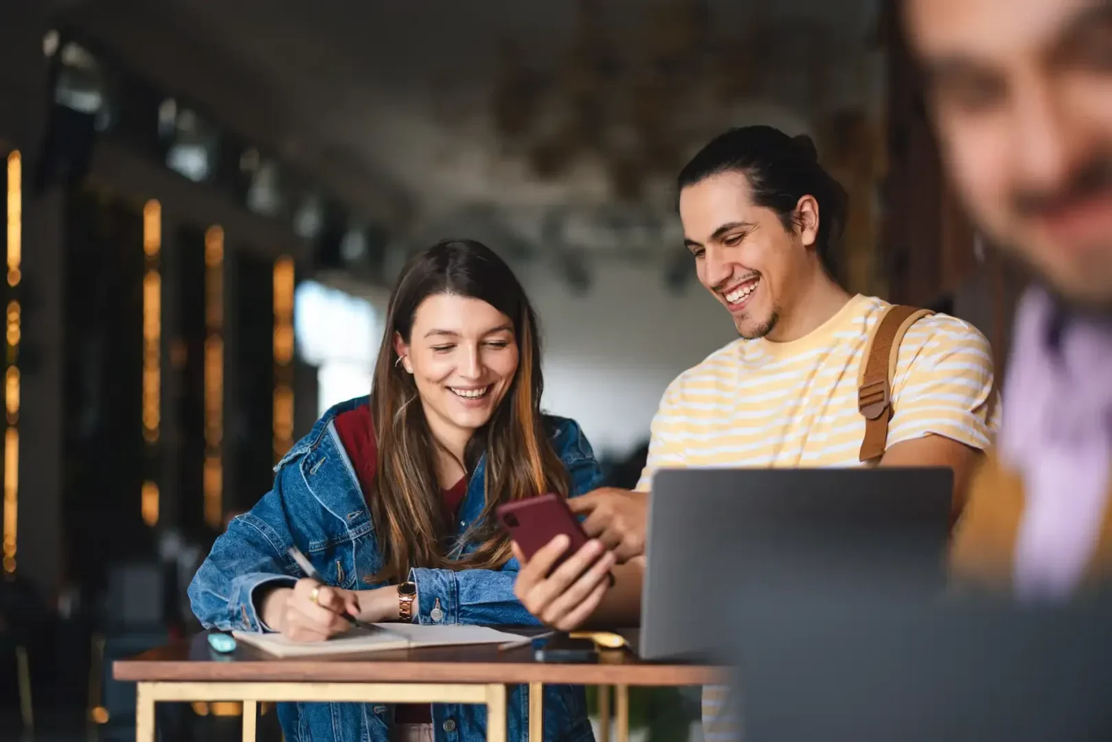Zwei junge Erwachsene lächeln, während sie gemeinsam auf ein Smartphone schauen. Der eine hält einen Stift und ein Notizbuch in der Hand, der andere arbeitet an einem Tisch in einer hellen, modernen Innenraumumgebung an einem Laptop.
