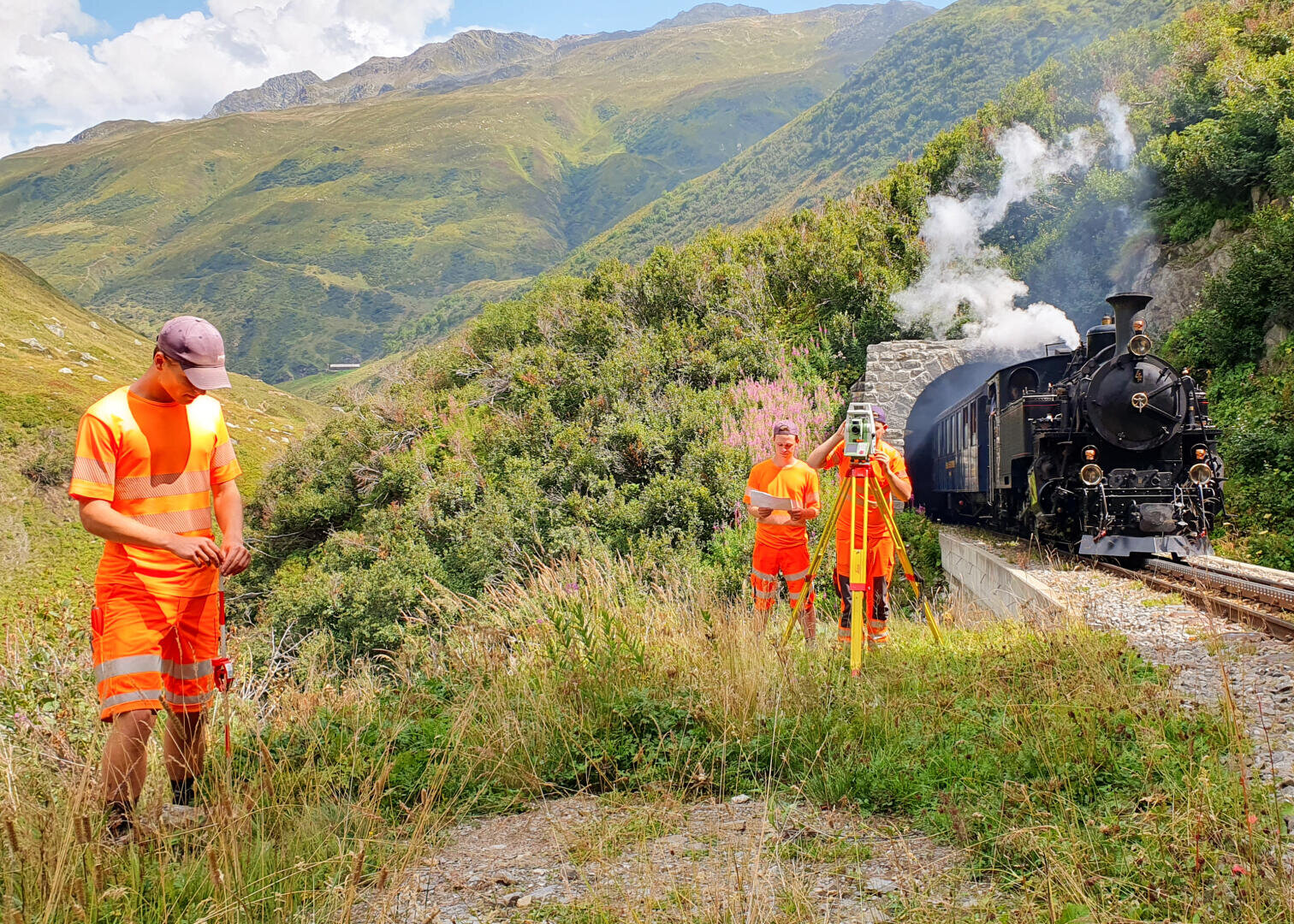 Drei Personen in orangefarbener Schutzkleidung arbeiten in einer grünen Berglandschaft in der Nähe von Bahngleisen. Ein schwarzer Dampfzug fährt aus einem Tunnel heraus, und eine Person bedient neben den Gleisen Vermessungsgeräte.