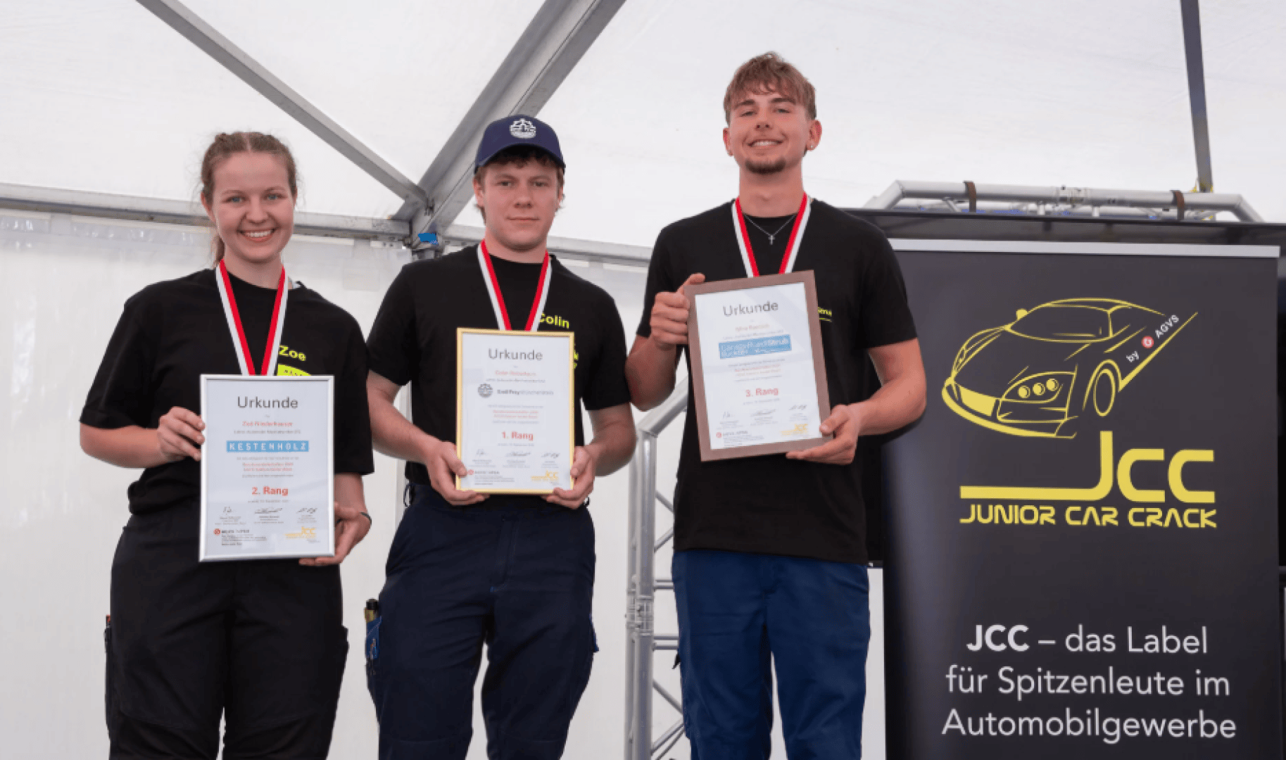 Drei junge Menschen, die Medaillen tragen und Urkunden in den Händen halten, stehen auf einem Podium. Sie lächeln. Auf einem Banner neben ihnen steht „JCC Junior Car Crack“ und ist mit einer gelben Autografik verziert. Die Szene scheint eine Preisverleihung zu sein.
