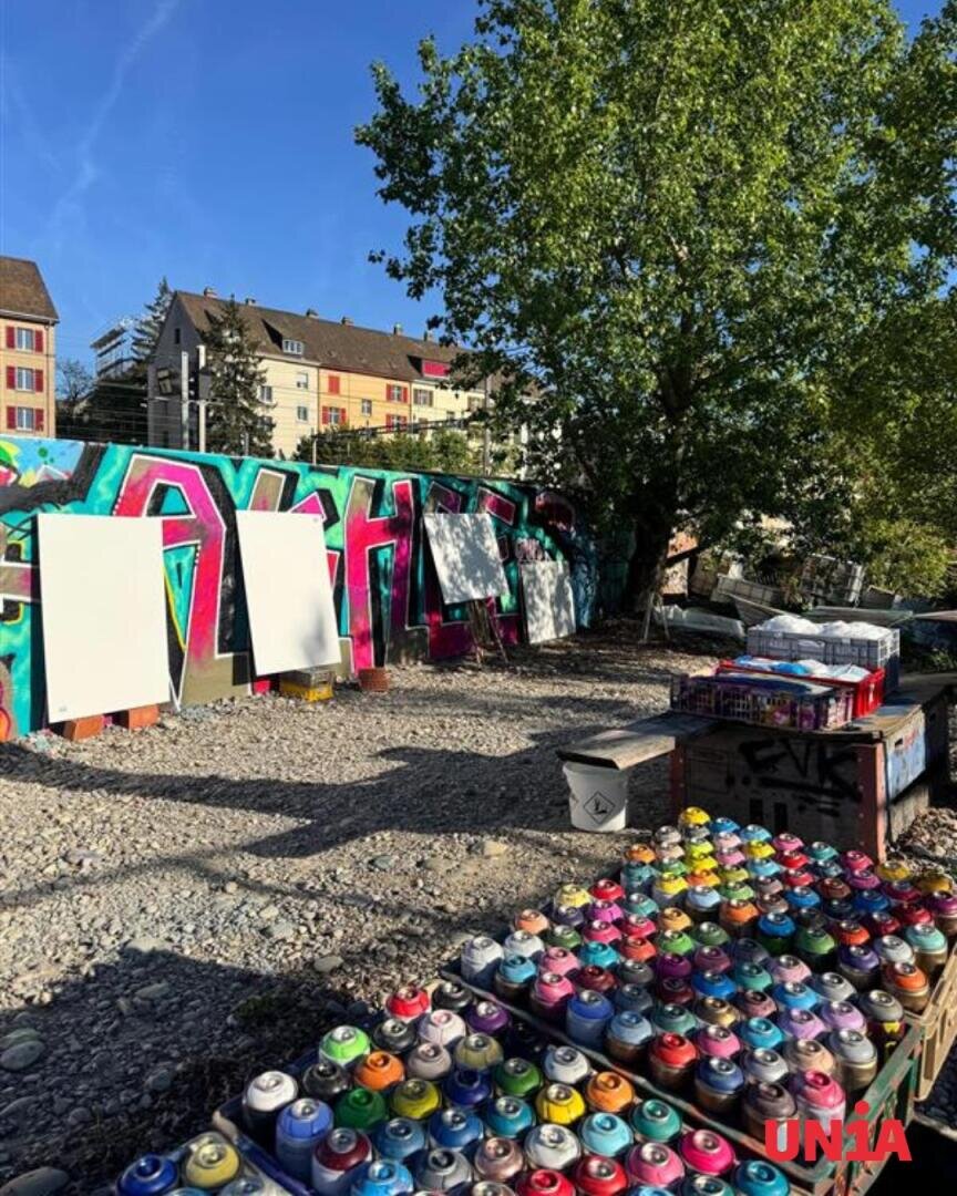 Auf einem Tisch vor leeren Leinwänden und einer bunten, mit Graffiti verzierten Wand stehen Sprühdosen, im Hintergrund sind Bäume und Gebäude vor einem strahlend blauen Himmel zu sehen.
