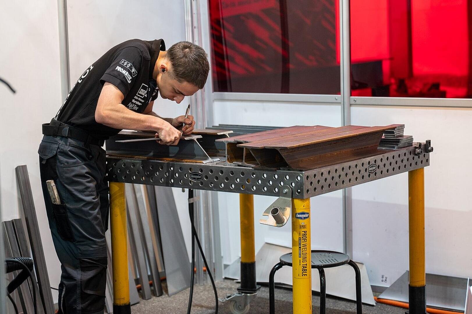 Eine Person in schwarzer Kleidung bearbeitet mit einem Werkzeug Metallteile auf einer gelochten Werkbank, im Hintergrund sind weitere Metallstangen und eine rote Trennwand zu sehen.