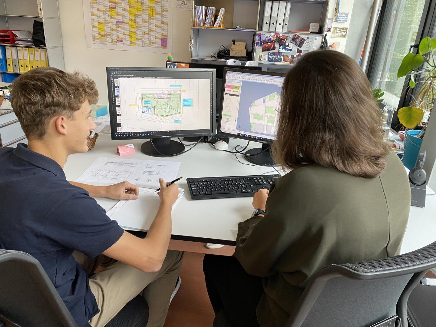 Zwei Personen sitzen an einem Schreibtisch und arbeiten an Architekturentwürfen, die auf zwei Monitoren angezeigt werden. Eine zeigt auf ein Blatt Papier, während die andere die Tastatur bedient. Der Arbeitsplatz ist mit Regalen, Pflanzen und Unterlagen ausgestattet.