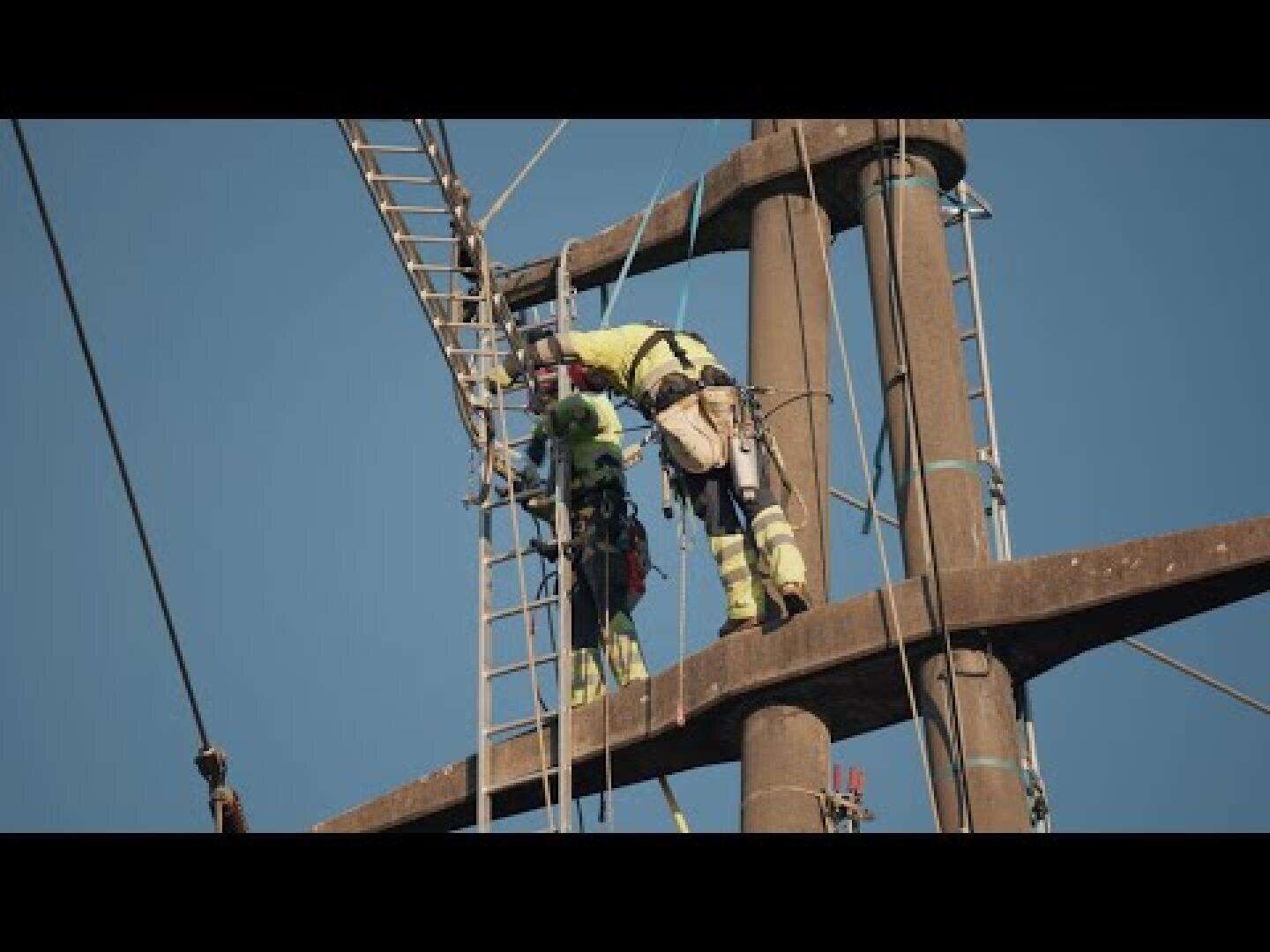 Zwei Arbeiter in Schutzkleidung stehen auf einem hohen Strommast und nutzen dabei Leitern und Sicherheitsgurte vor dem Hintergrund eines strahlend blauen Himmels.