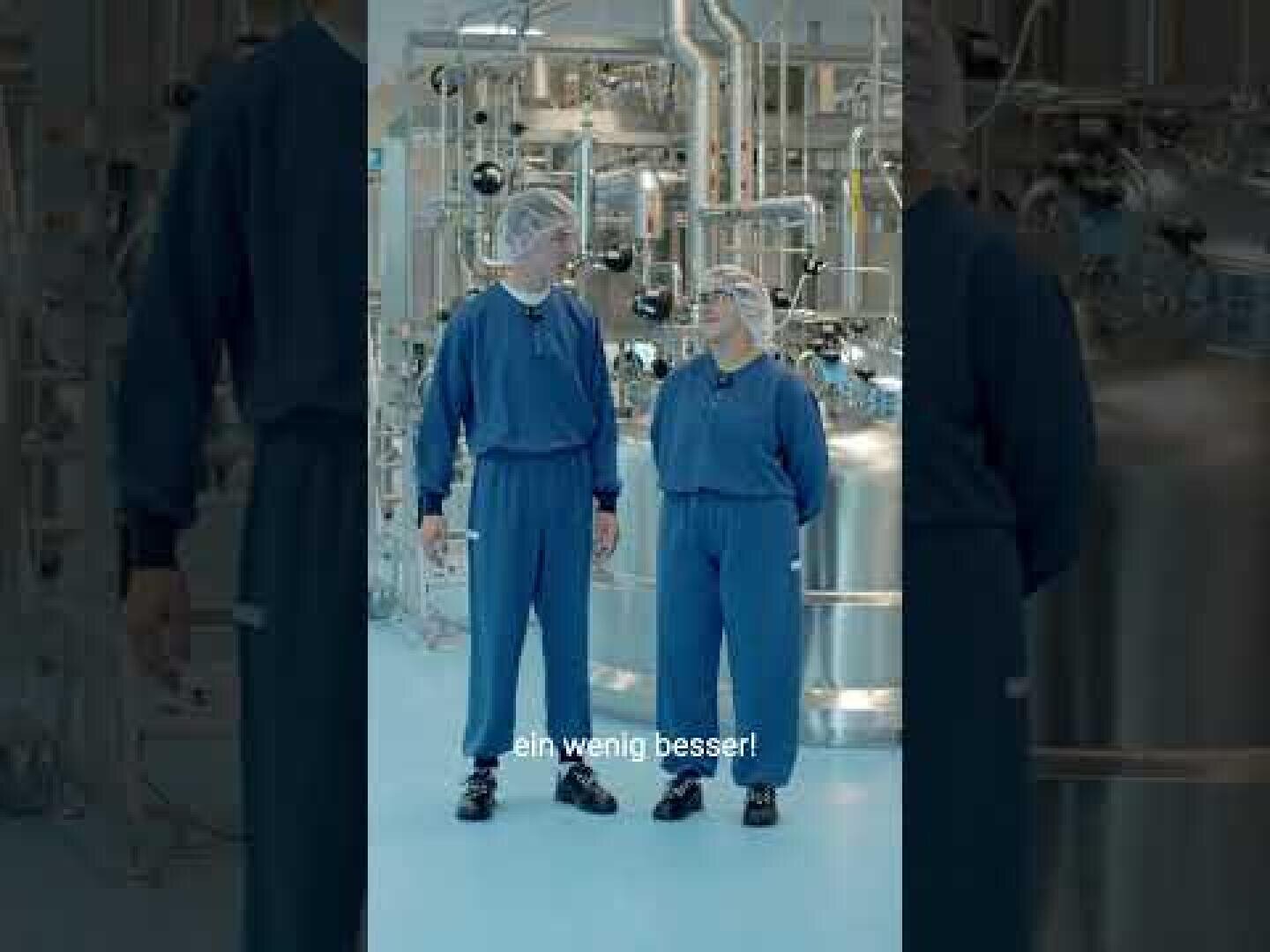 Zwei Personen in blauen Schutzanzügen und Haarnetzen stehen in einem hell beleuchteten Industrielabor, im Hintergrund sind Metallrohre und Geräte zu sehen. Unten im Bild ist ein weißer Untertitel zu lesen: „ein wenig besser!“.