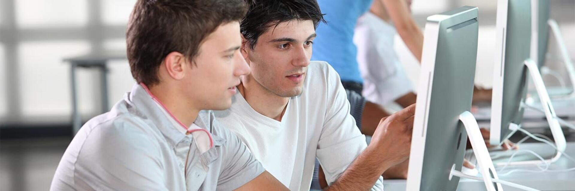 Zwei junge Männer sitzen nebeneinander und blicken auf den Bildschirm eines Desktop-Computers, wobei einer mit dem Finger auf den Monitor zeigt. Sie wirken konzentriert und engagiert und arbeiten möglicherweise gemeinsam an einem Projekt in einem modernen, hellen Raum, in dessen Hintergrund weitere Personen zu sehen sind.