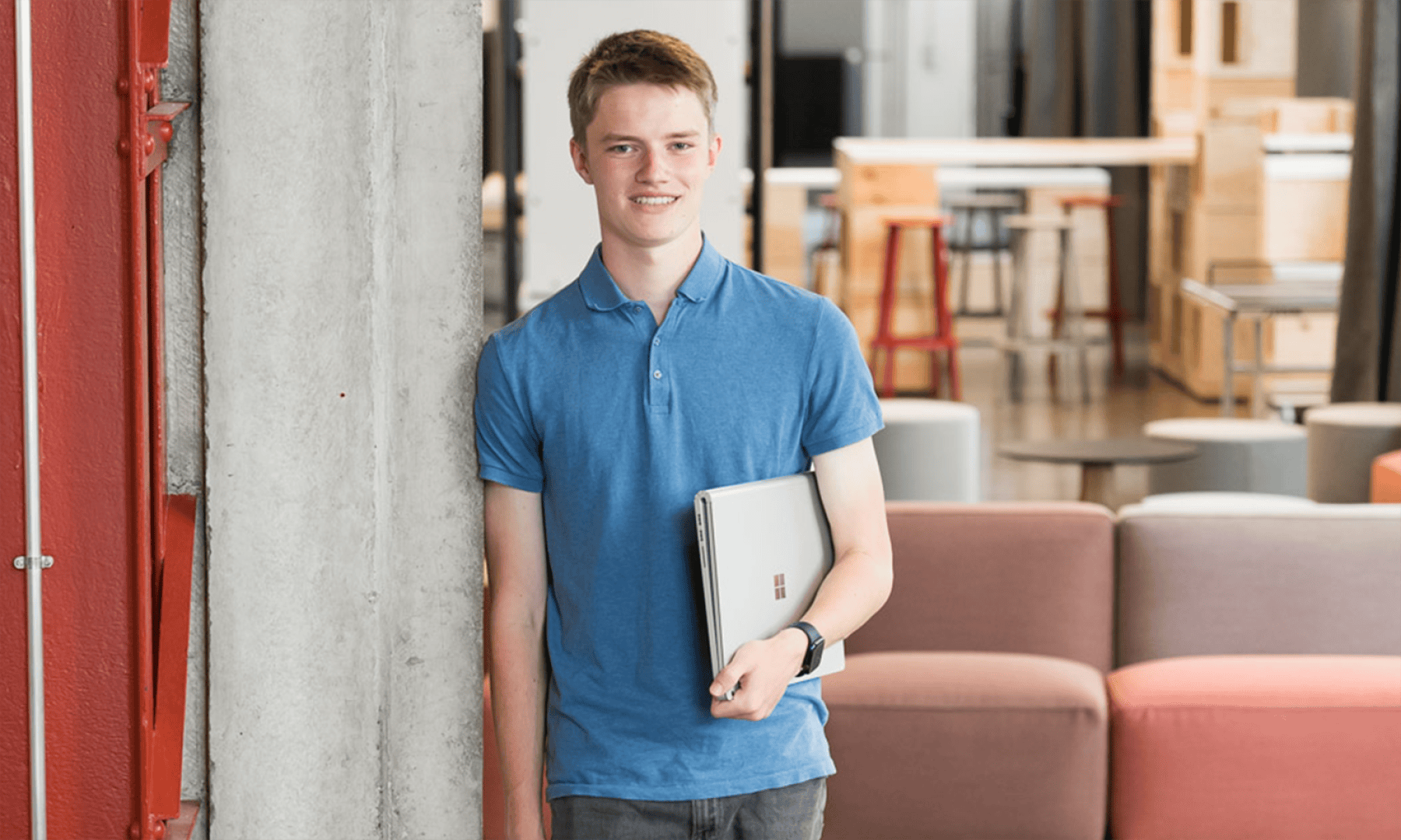 Ein junger Mensch in einem blauen Polohemd steht in einem Raum, lächelt und hält einen geschlossenen Laptop in der Hand. Im Hintergrund sind moderne Büromöbel zu sehen, darunter Sofas und Hocker.
