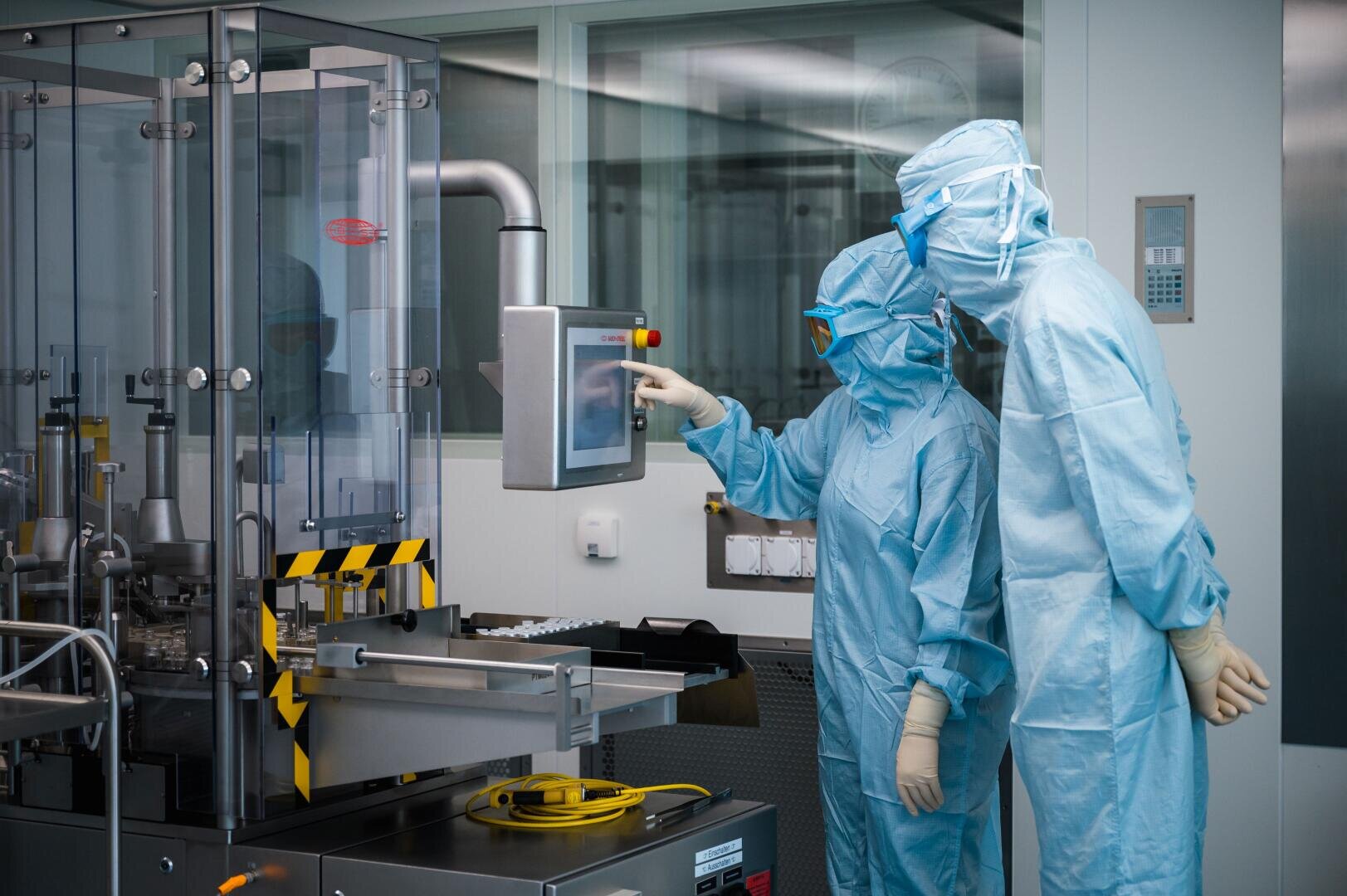 Zwei Personen in blauen Schutzanzügen bedienen einen Touchscreen an einer Maschine in einem sterilen Labor, umgeben von sauberen Geräten und Glaswänden.