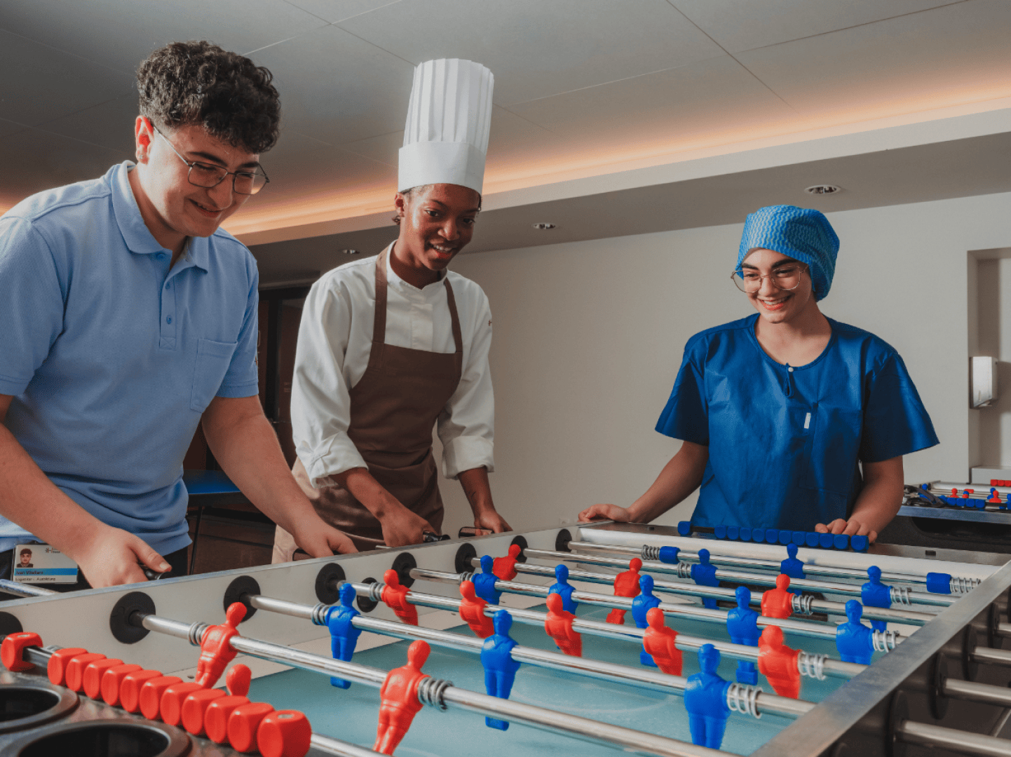Drei Personen – eine mit Kochmütze, eine in OP-Kleidung und eine im Polohemd – spielen fröhlich gemeinsam drinnen Tischfußball.