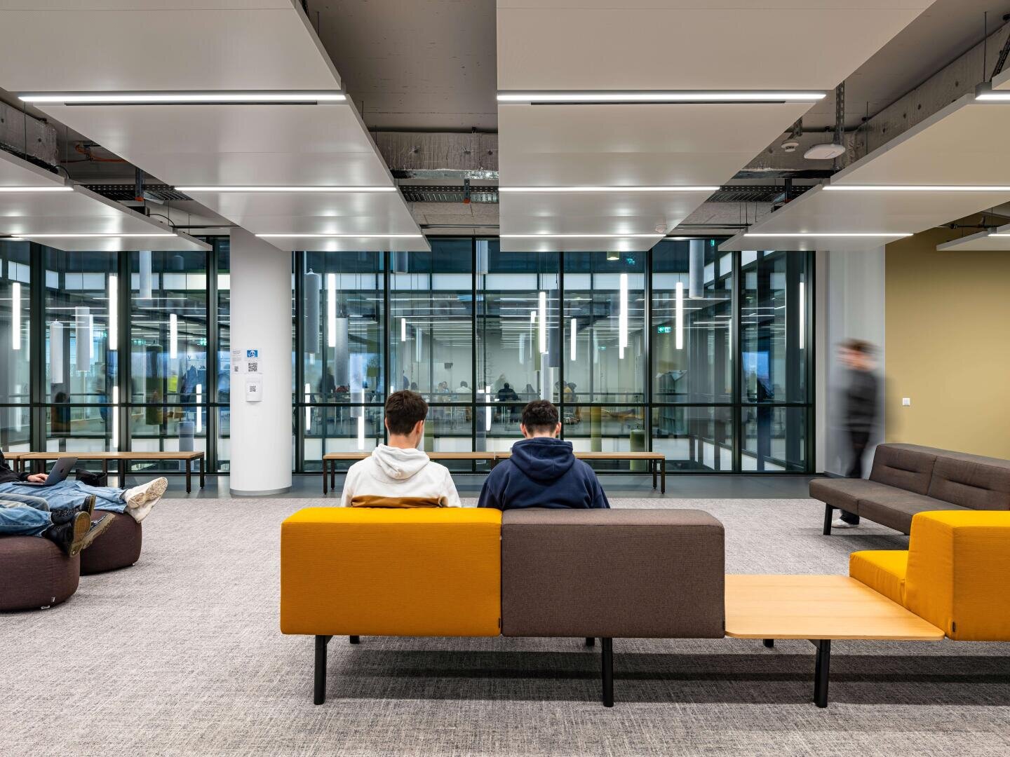 Zwei Personen sitzen auf einem gelb-braunen Sofa in einem modernen, offenen Wohnbereich mit großen Fenstern, heller Beleuchtung und vereinzelten Sitzgelegenheiten. Im Hintergrund sind weitere Personen zu sehen.