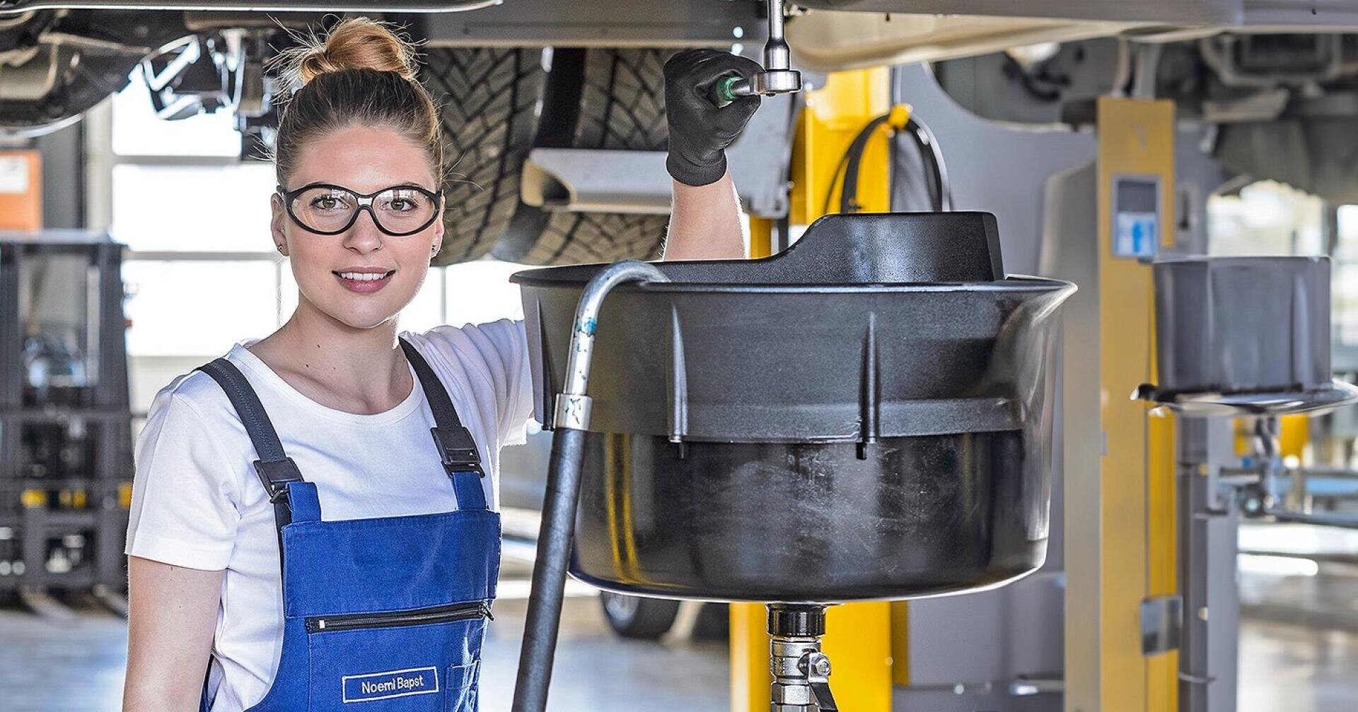 Eine Frau mit Brille und blauer Latzhose steht in einer Autowerkstatt und lächelt, während sie mit professionellem Werkzeug und professioneller Ausrüstung Wartungsarbeiten unter einem Fahrzeug durchführt.