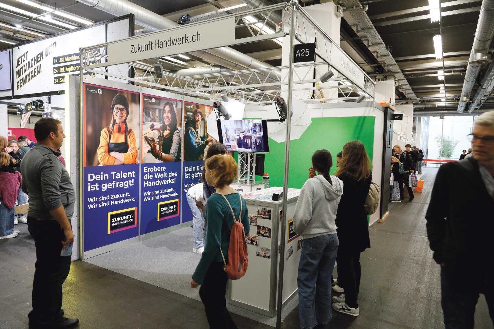 Besucher besichtigen den Stand von ZukunftHandwerk.ch auf einer Indoor-Veranstaltung. Bunte Plakate werben für Berufe im Handwerk. Die Besucher interagieren mit den Ausstellungsstücken und sehen sich die Informationen an, während andere in der Messehalle vorbeigehen.
