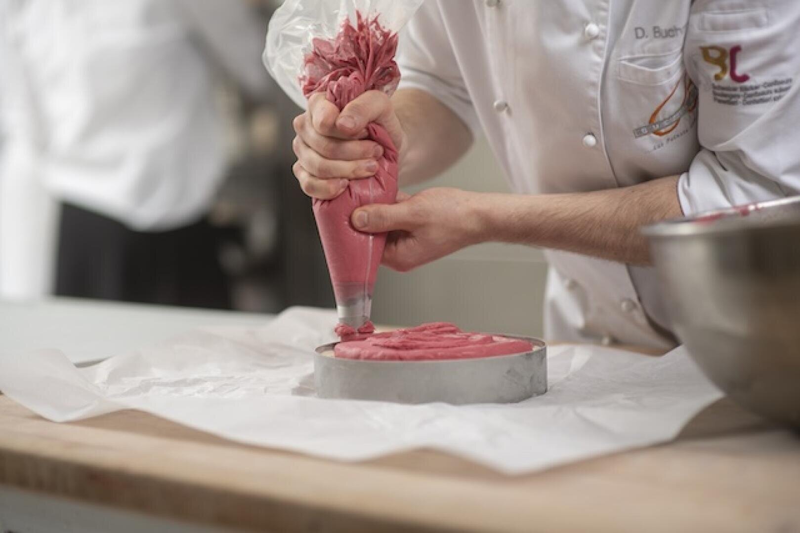 Ein Koch in weißer Kochkleidung spritzt mit einem Spritzbeutel rosa Teig oder Zuckerguss in eine runde Kuchenform, die auf einer mit Backpapier ausgelegten Arbeitsfläche steht. Daneben steht eine Metallschüssel auf der Holzarbeitsplatte.