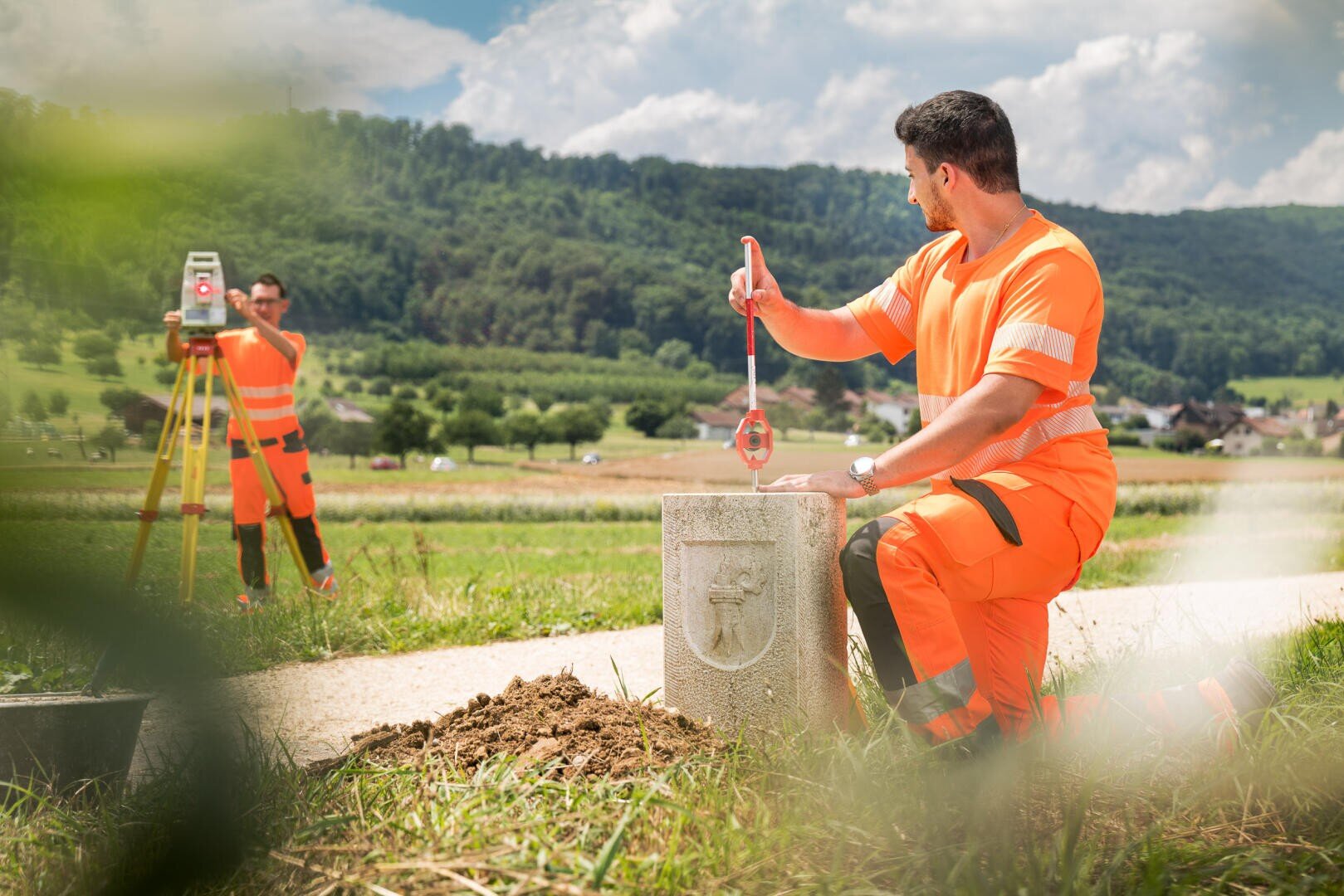 Zwei Arbeiter in orangefarbener Schutzkleidung vermessen und markieren ein Grundstück entlang eines ländlichen Weges; einer misst mit einem Maßband an einem Steinmarker ab, während der andere Vermessungsgeräte bedient. Im Hintergrund sind grüne Felder und Hügel zu sehen.