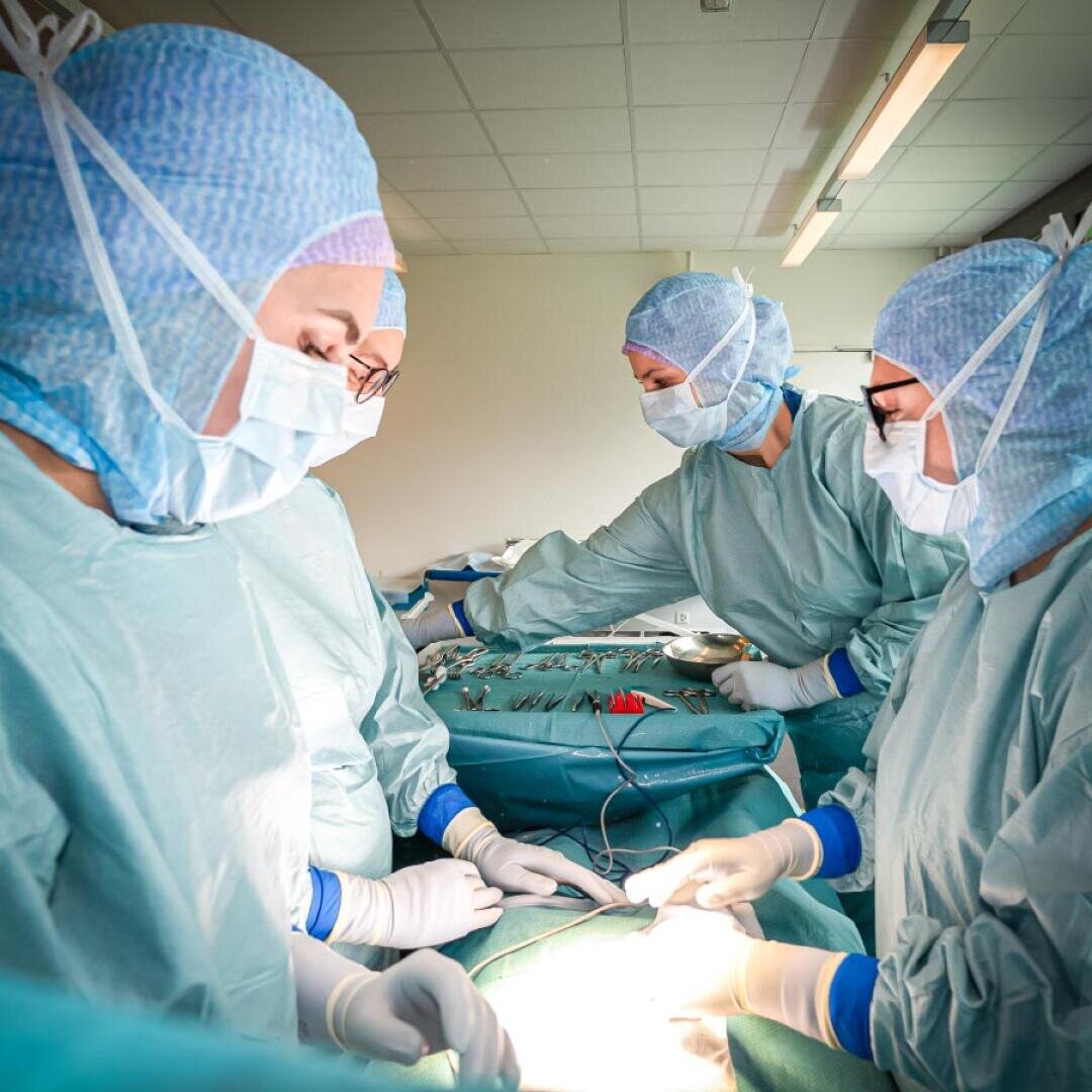 Vier Chirurgen in blauen OP-Kitteln und Masken operieren in einem hell erleuchteten Krankenhausraum und konzentrieren sich auf einen Patienten, während medizinische Instrumente auf einem Tisch in der Nähe bereitliegen.