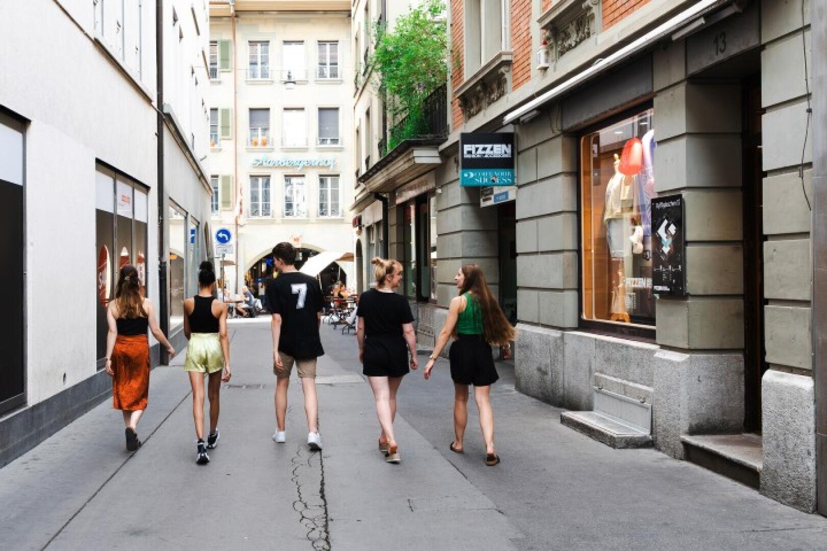 An einem sonnigen Tag gehen fünf junge Leute eine schmale Stadtstraße entlang, die von Geschäften und Wohnungen gesäumt ist. Vier von ihnen halten sich paarweise an den Händen, während einer einen Regenschirm hält. Im Hintergrund sitzen andere Leute in einem Café.