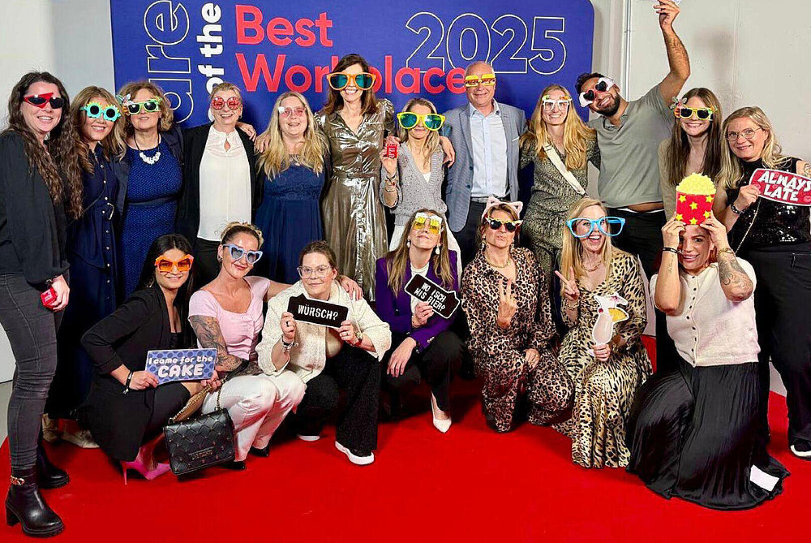Eine Gruppe von Menschen posiert auf einem roten Teppich vor dem Hintergrund der "Best Workplaces 2025". Sie tragen lustige Brillen und halten verspielte Requisiten in der Hand, lächeln und feiern gemeinsam.