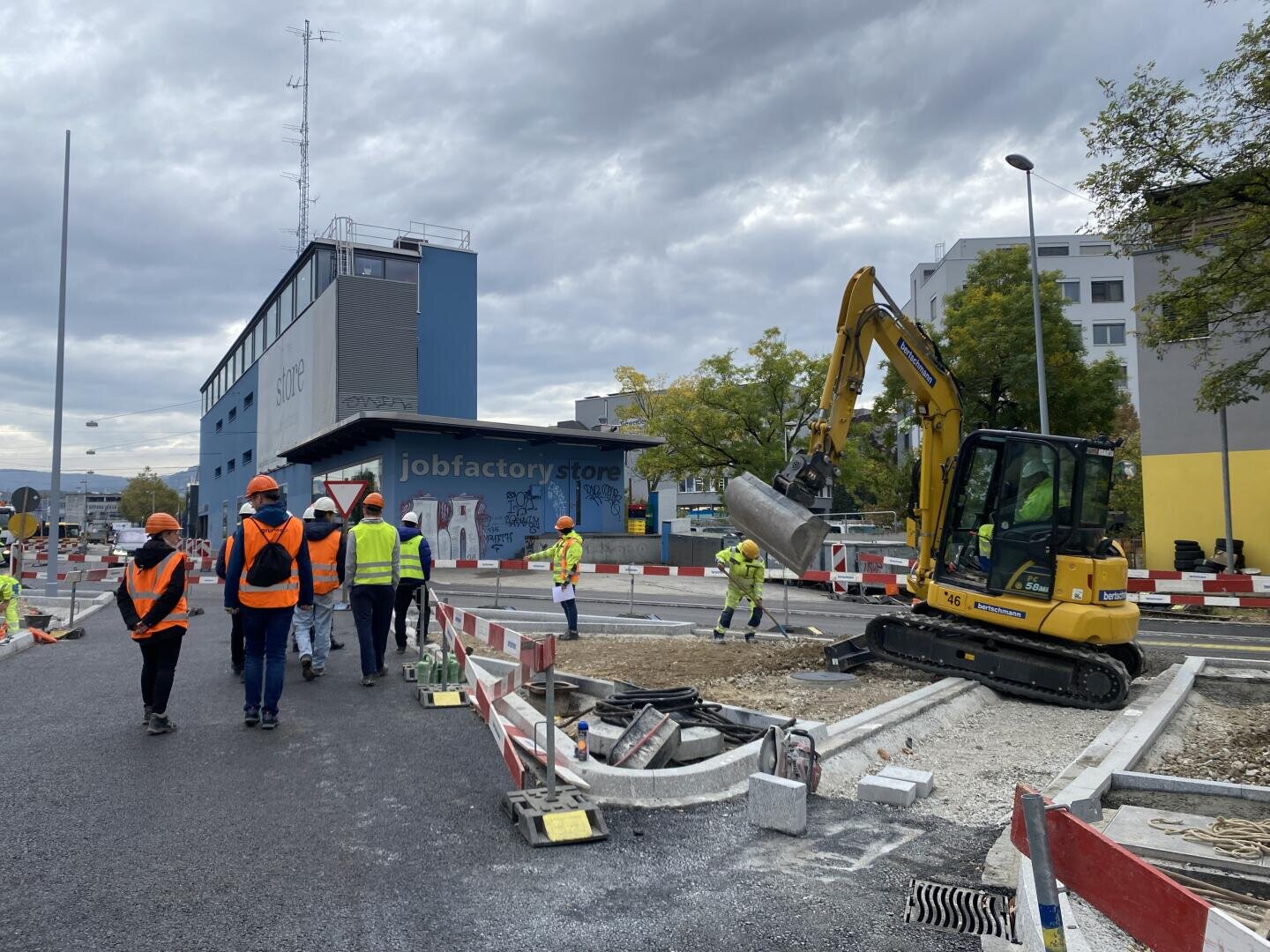 Bauarbeiter in Sicherheitswesten und mit Helmen arbeiten in der Nähe eines gelben Baggers auf einer Straße in der Stadt. Im Hintergrund sind Absperrungen, Leitkegel und Baumaterialien zu sehen, vor einem bewölkten Himmel und Gebäuden.