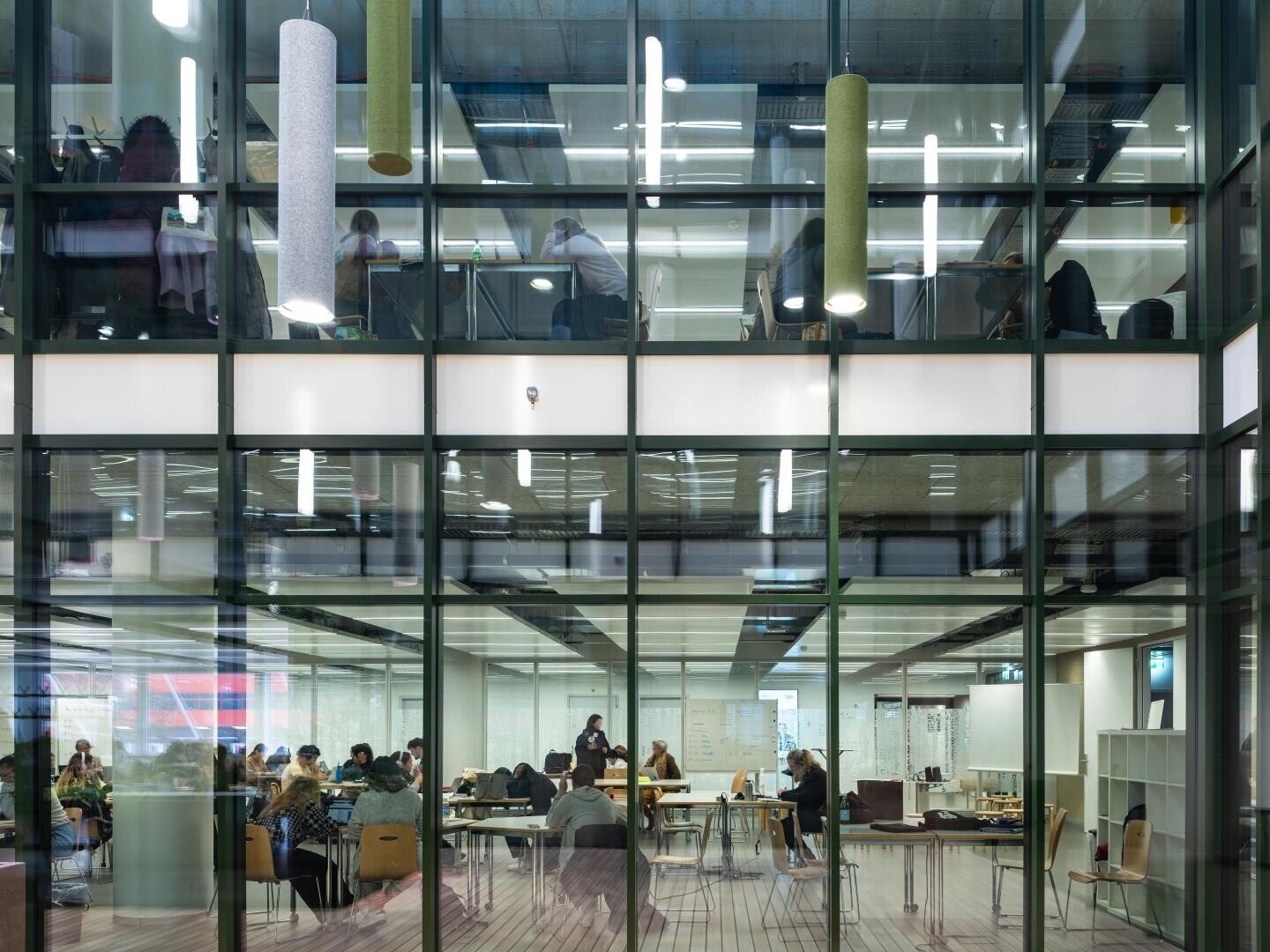 In einer modernen, glasverglasten Bibliothek oder einem Bürogebäude mit zwei sichtbaren Stockwerken und heller Beleuchtung sitzen Menschen an Tischen und lernen oder arbeiten.