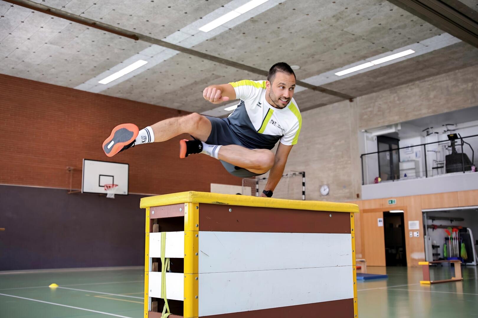 Ein Mann in Sportkleidung springt in einer Sporthalle über eine Sprungbox und stützt sich dabei mit einer Hand ab. Im Hintergrund sind Basketballkörbe und Turngeräte zu sehen.