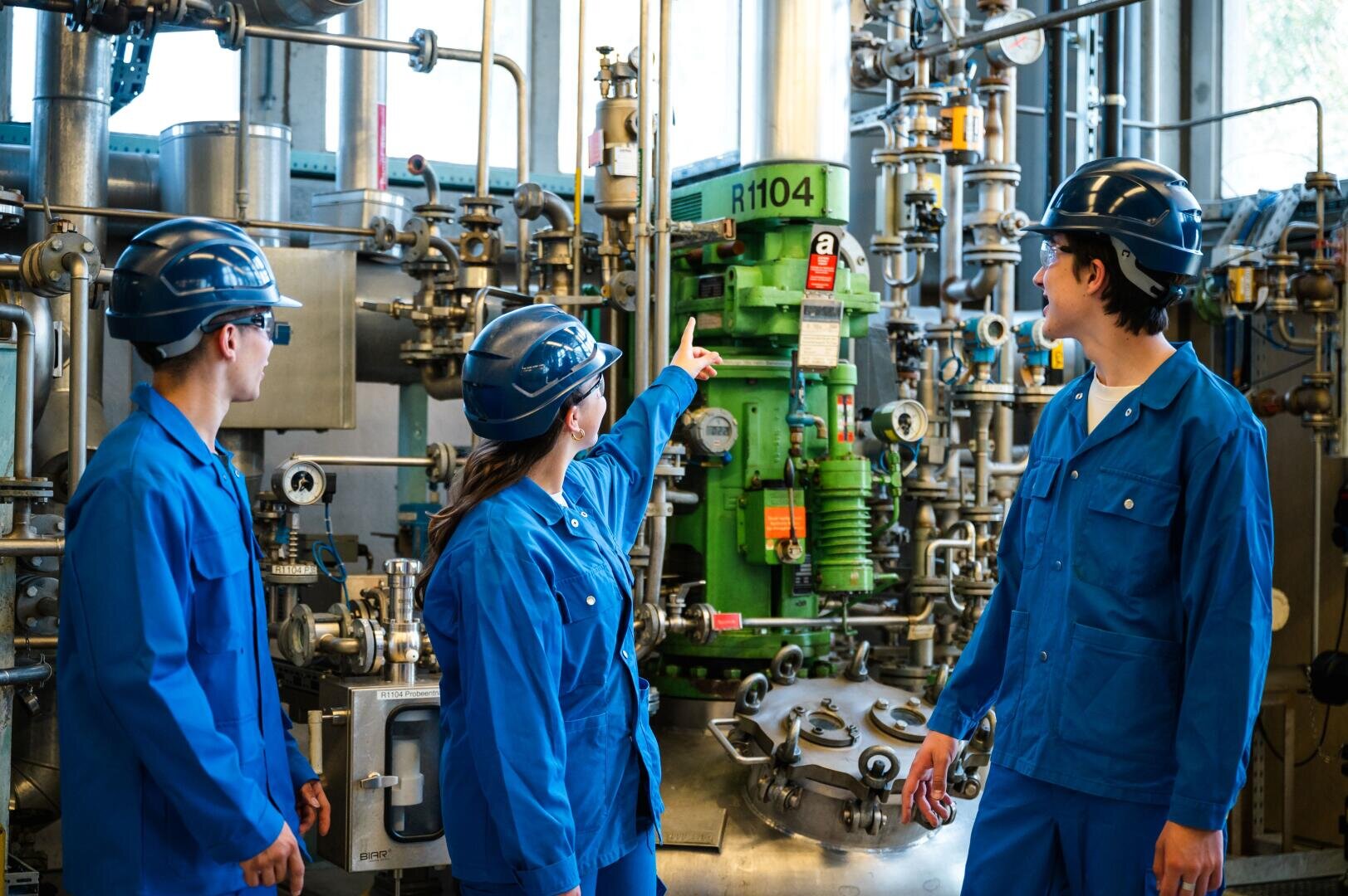 Drei Arbeiter in blauen Overalls und mit Schutzhelmen stehen in einer Industrieanlage mit Rohrleitungen und Tanks. Ein Arbeiter zeigt auf ein grünes Bauteil mit der Aufschrift „R1104“, während die anderen in dieselbe Richtung blicken.
